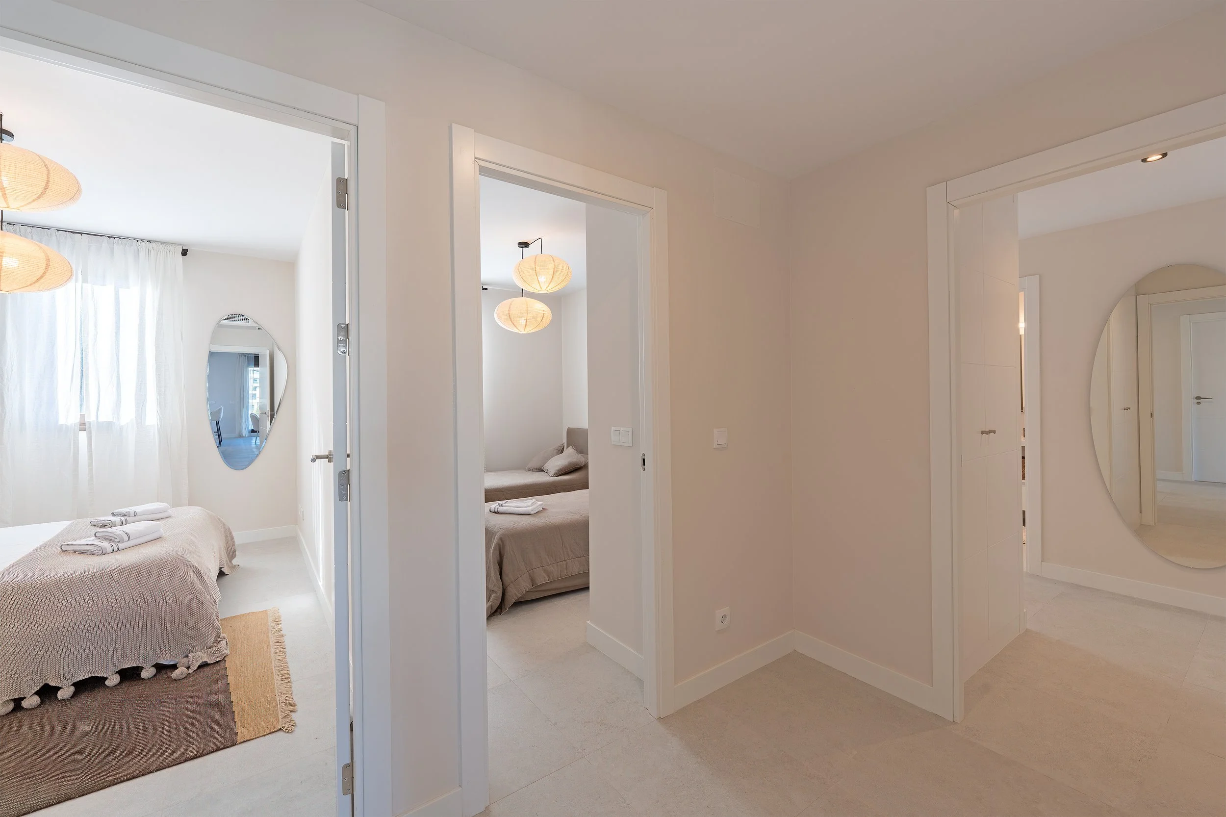View of a bright, modern apartment with two bedrooms, neutral-colored walls, and hanging lamps. One bedroom has a bed with towels on it, and there's an oval mirror on the wall.