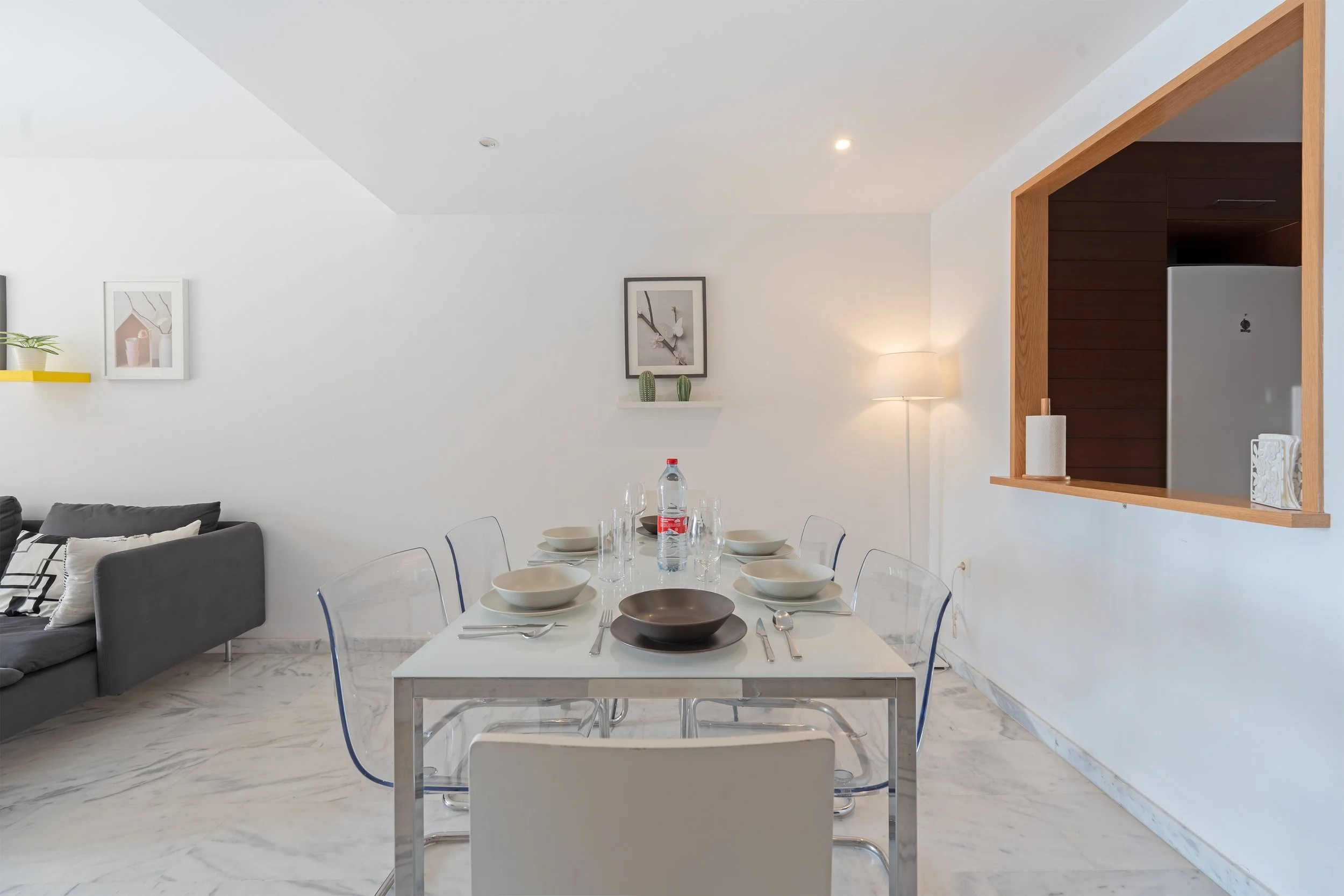 Dining room with a white table set with bowls, glasses, and cutlery, six clear chairs, a gray sofa with pillows, a wall-mounted shelf with framed art and small cactus plants, a floor lamp, and a window opening in the wall showing part of a refrigerat