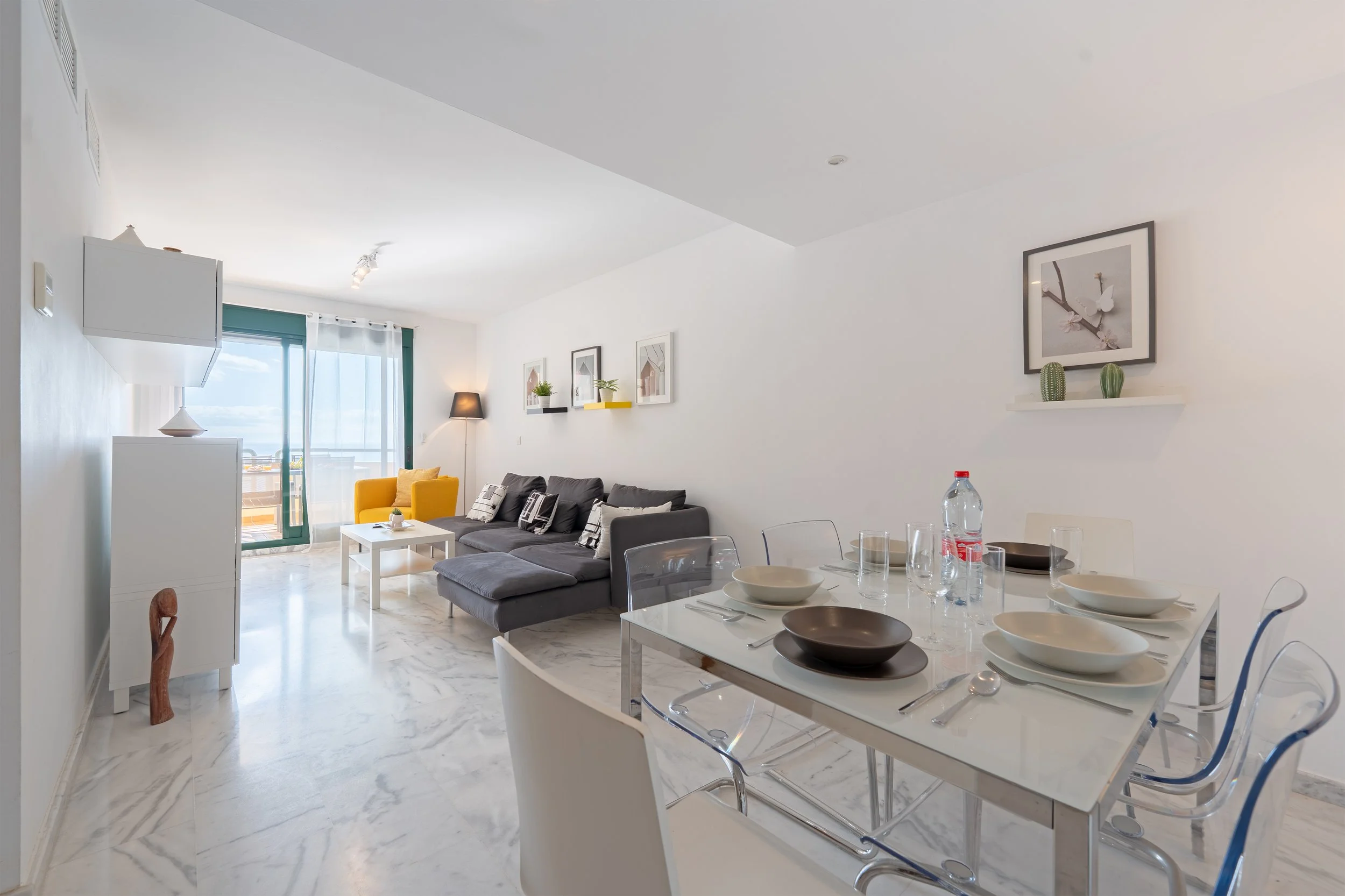 Modern living room and dining area with white walls, marble flooring, a gray sectional sofa, a yellow armchair, a white coffee table, a dining table with six chairs, and a balcony with sliding glass doors.