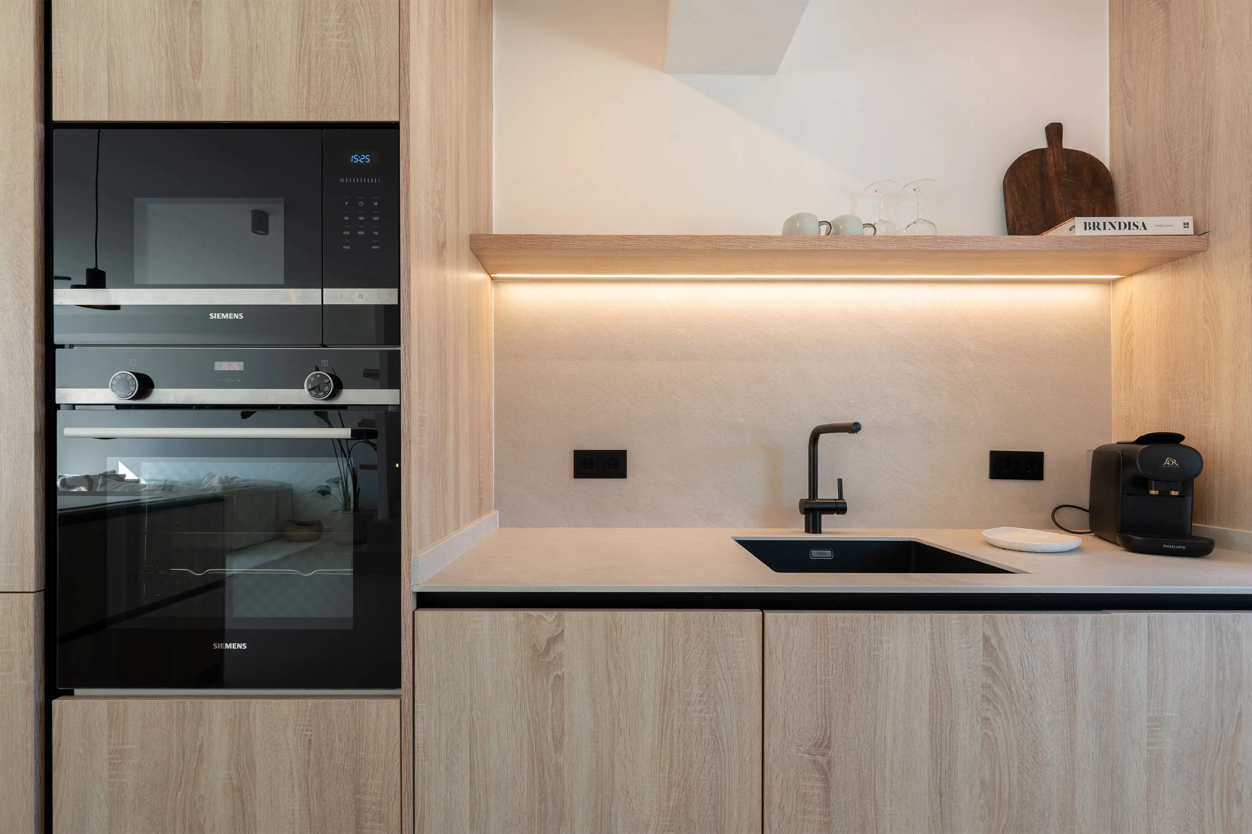 Modern kitchen with built-in Siemens oven and microwave, black sink, black faucet, coffee machine, and wooden cabinetry with a shelf holding cups, glasses, cutting board, and a book.