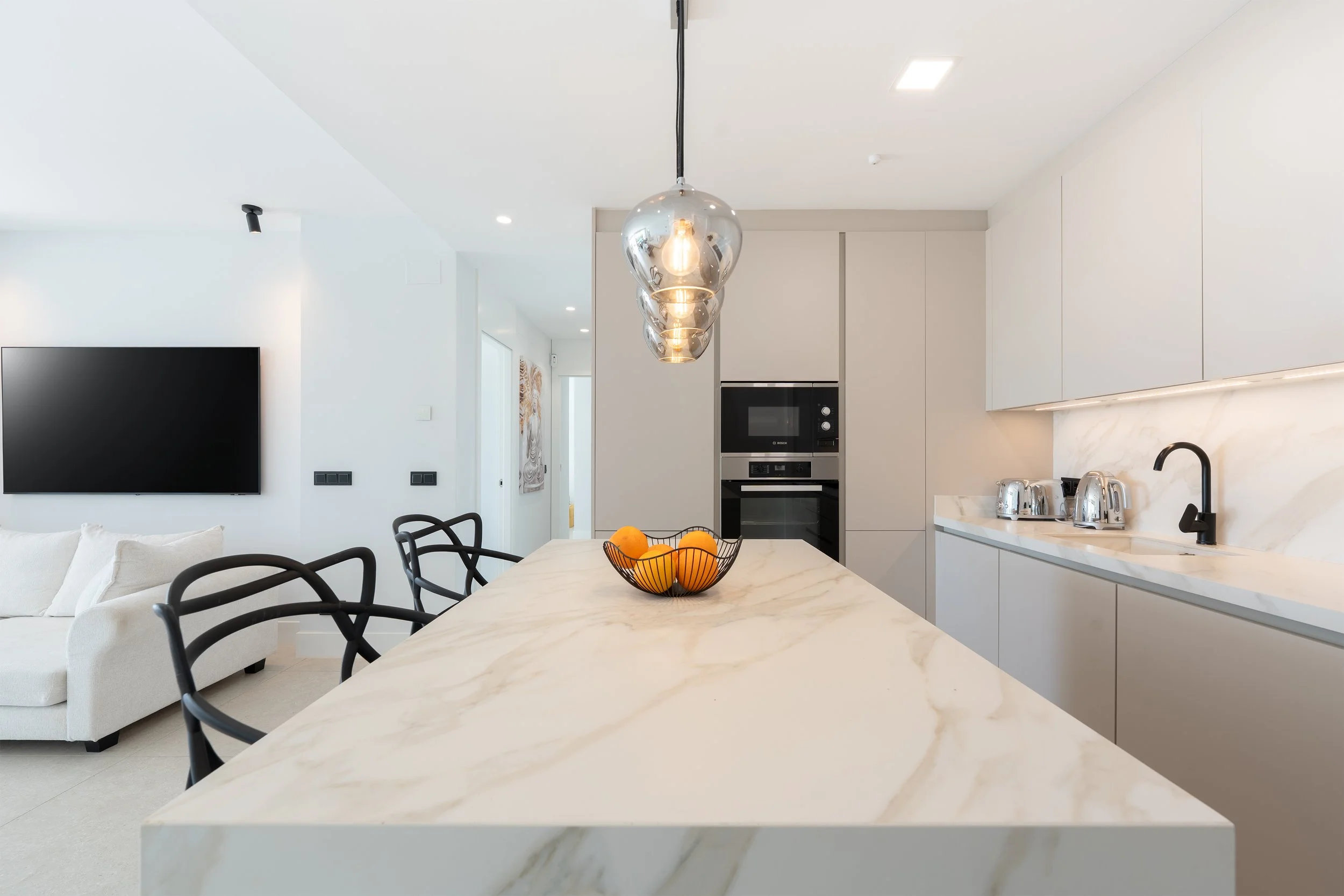 Modern open-concept kitchen with white marble island, black chairs, pendant lighting, and built-in appliances.