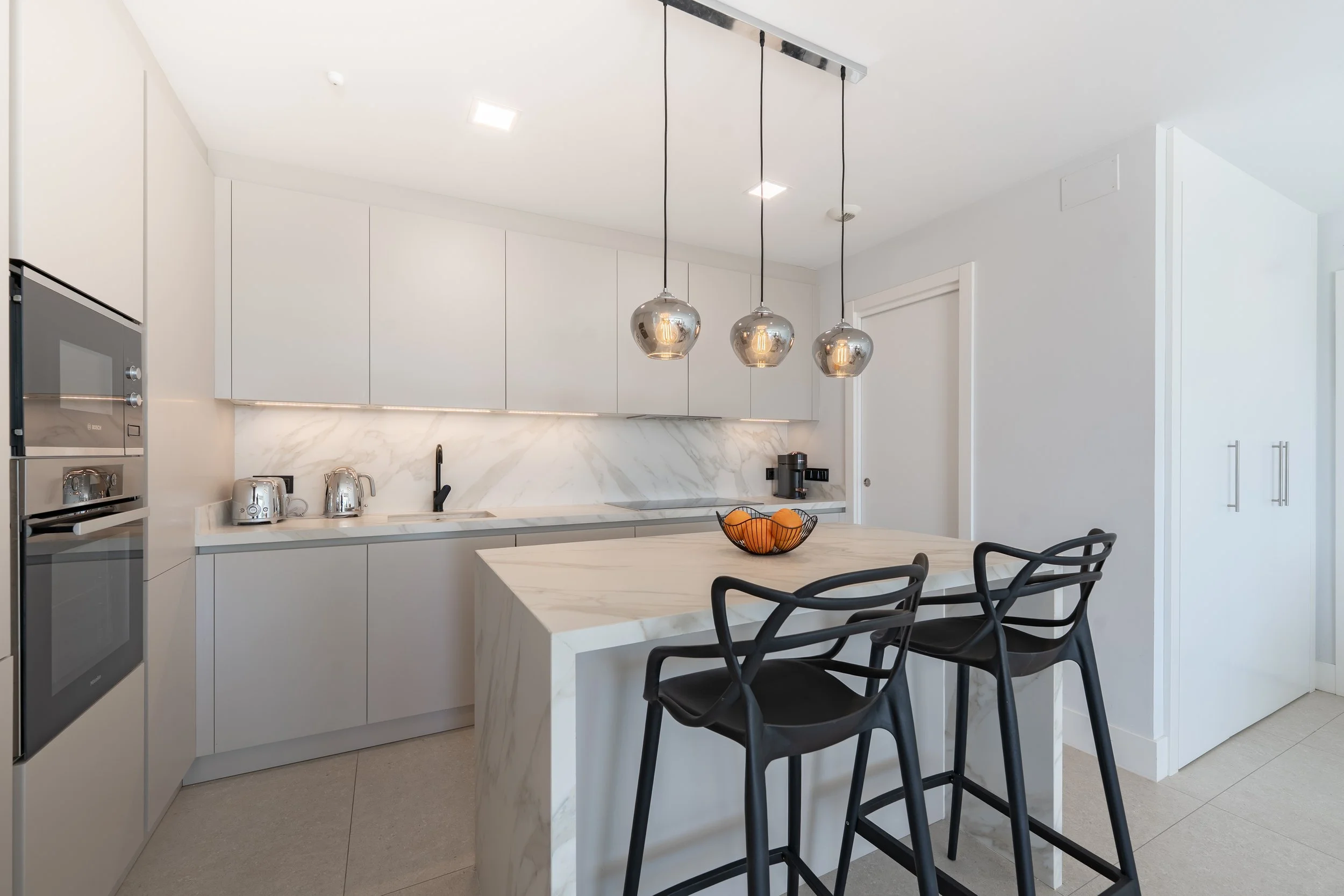 Modern, minimalist kitchen with white cabinetry, marble countertops and backsplash, and black accents. Features a central island with bar stools, pendant lighting, and kitchen appliances.