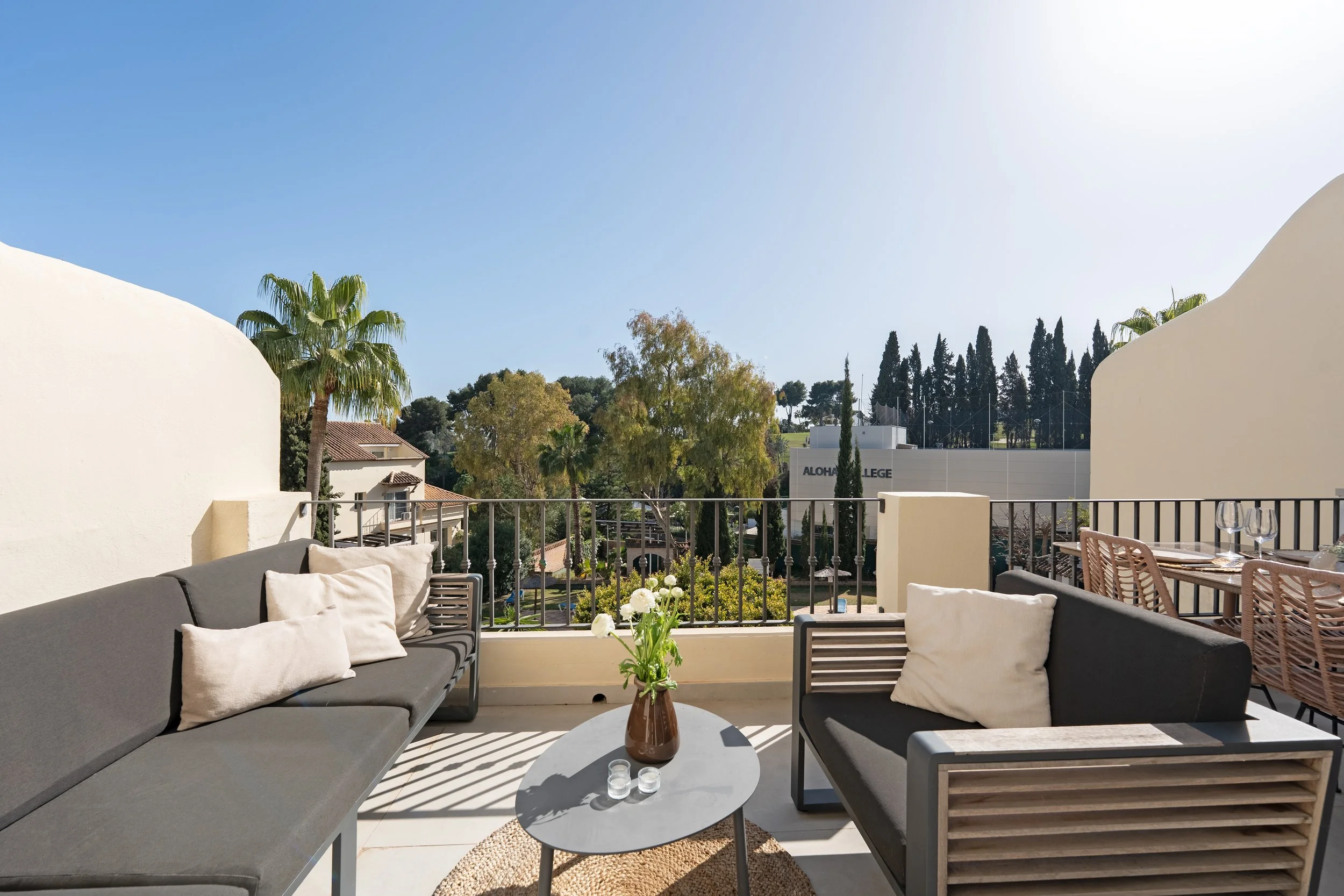 Outdoor balcony patio with seating area, two black sofas with white pillows, a small round table with a vase of white flowers and candles, surrounded by a black metal railing, overlooking trees and a building with the sign "Aloha College" in the dist