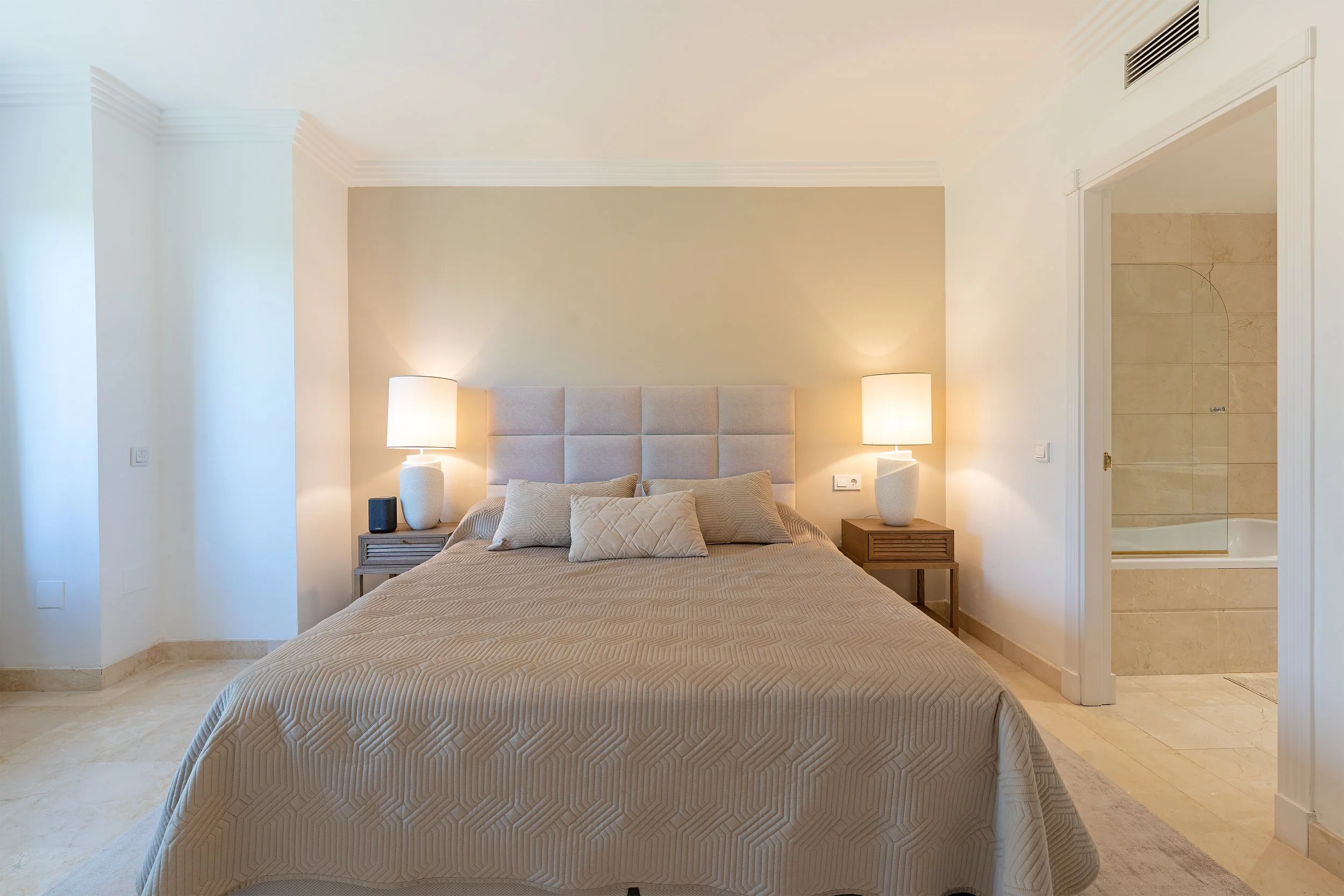 A bedroom with a king-size bed, beige quilted bedspread, four pillows, two matching nightstands with lamps, and an open doorway to a bathroom with a bathtub.
