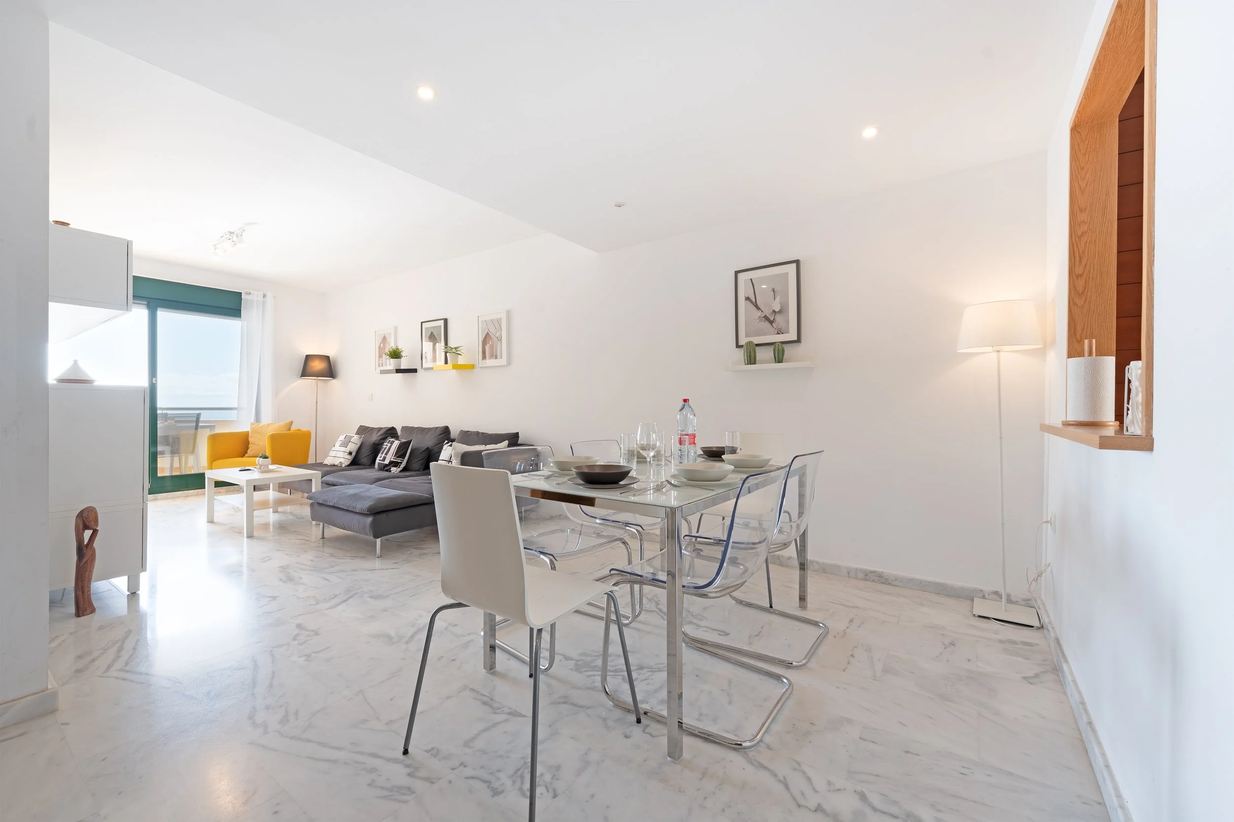 Living and dining area with white walls, marble floor, glass dining table with chairs, gray sectional sofa with pillows, yellow armchair, artwork on wall, floor lamps, sliding glass door leading to balcony.