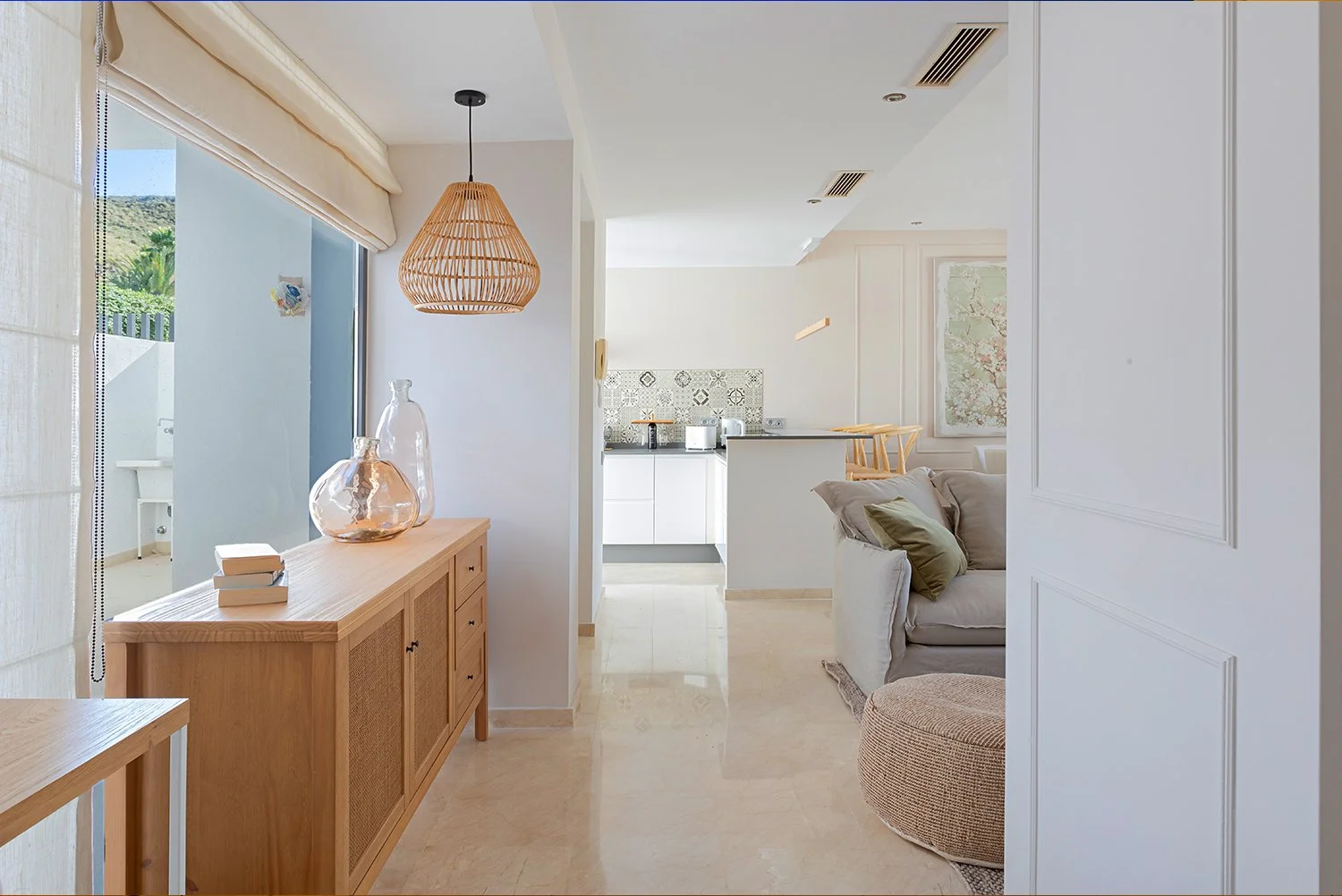 Interior of a modern, sunlit living space with light-colored walls, a wooden sideboard with decorative glass vases, a beige cushioned ottoman, and a sofa with green and beige cushions. In the background, a kitchen with patterned tiles and white cabin