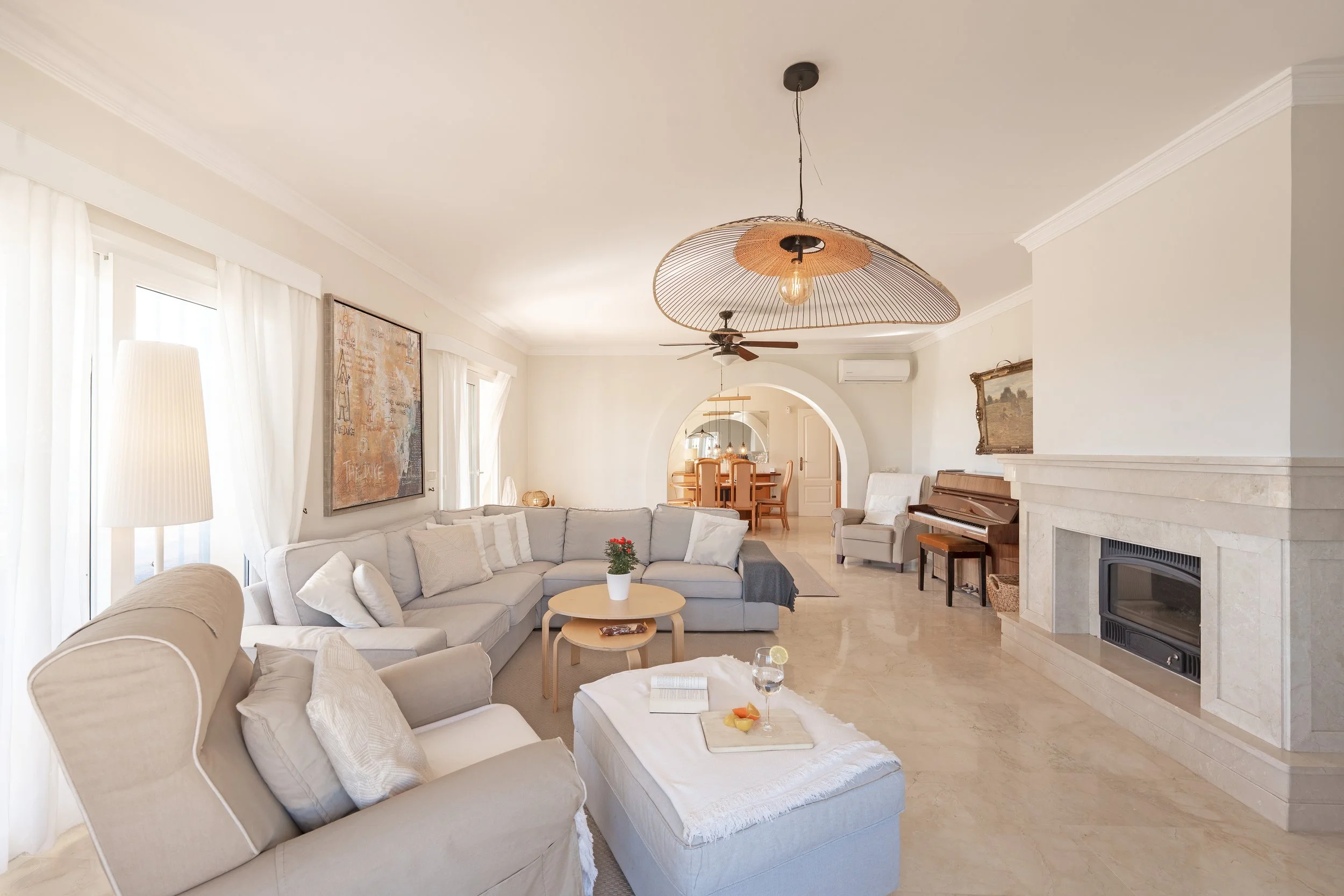 Bright living room with white sofa, beige armchair, potted plant, fireplace, hanging light fixture, ceiling fan, and view into dining area.