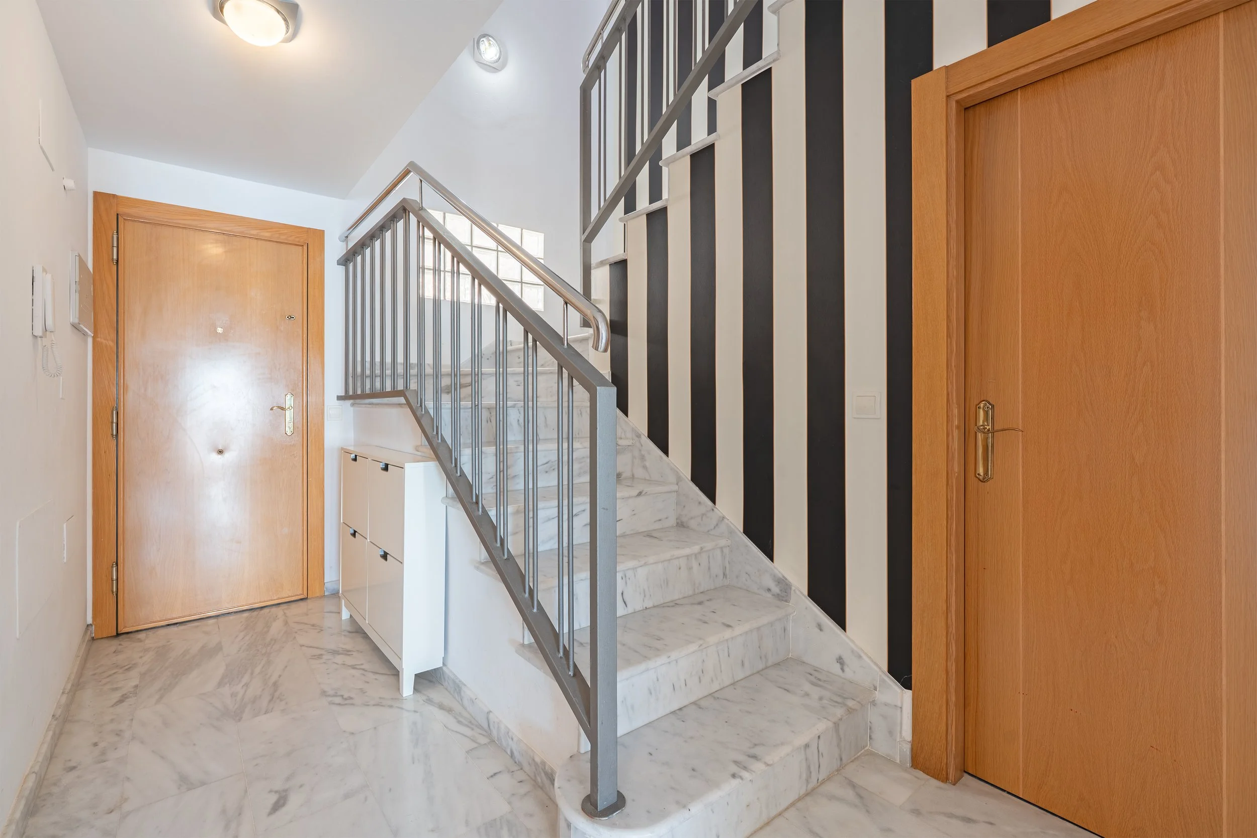 Interior of an entryway with marble flooring and staircase, featuring a wooden front door on the left, a wooden door on the right, a white cabinet under the staircase, and black and white vertical striped walls.