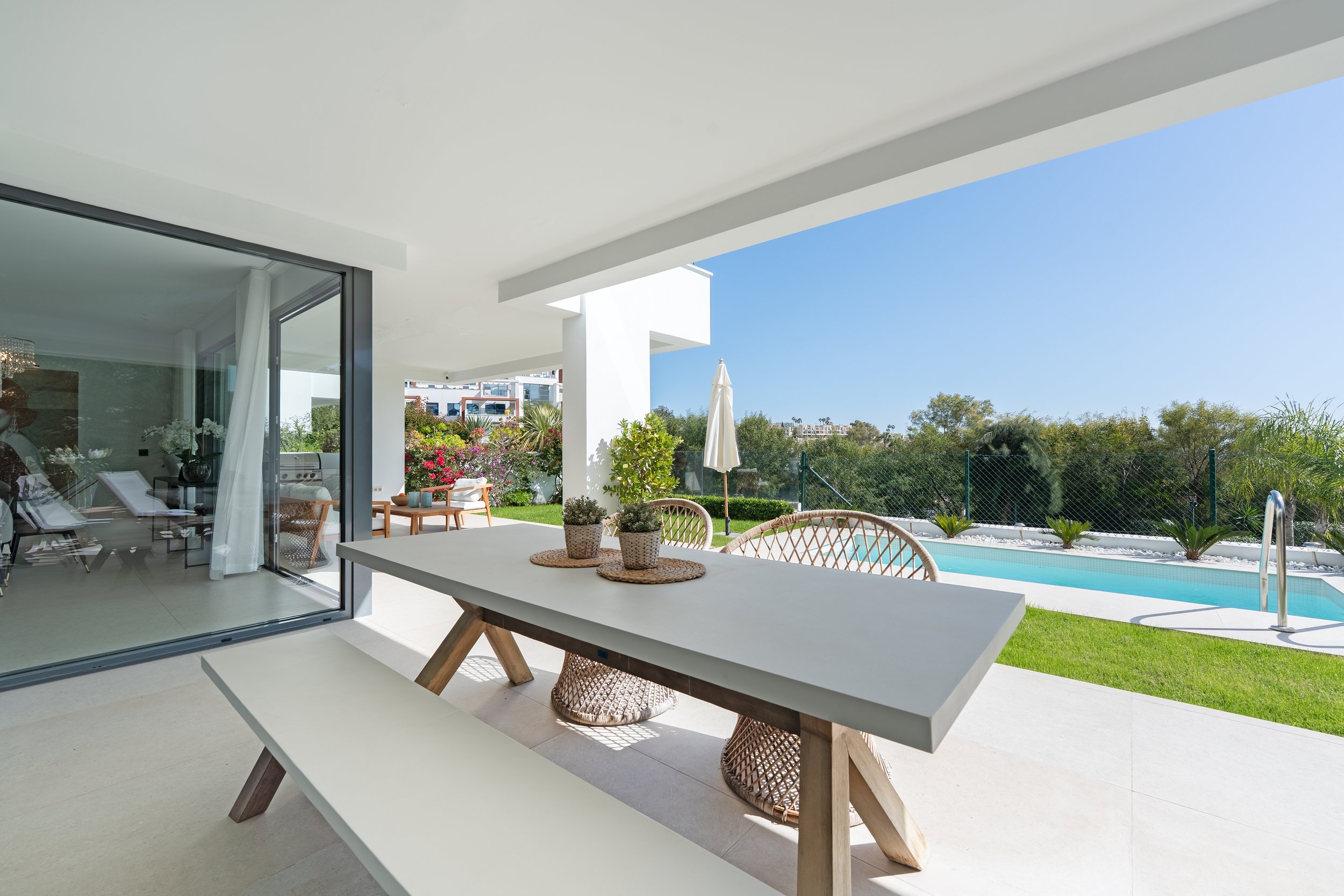 Modern patio with white table, wicker chairs, potted plants, overlooking a pool and lush greenery under clear blue sky.