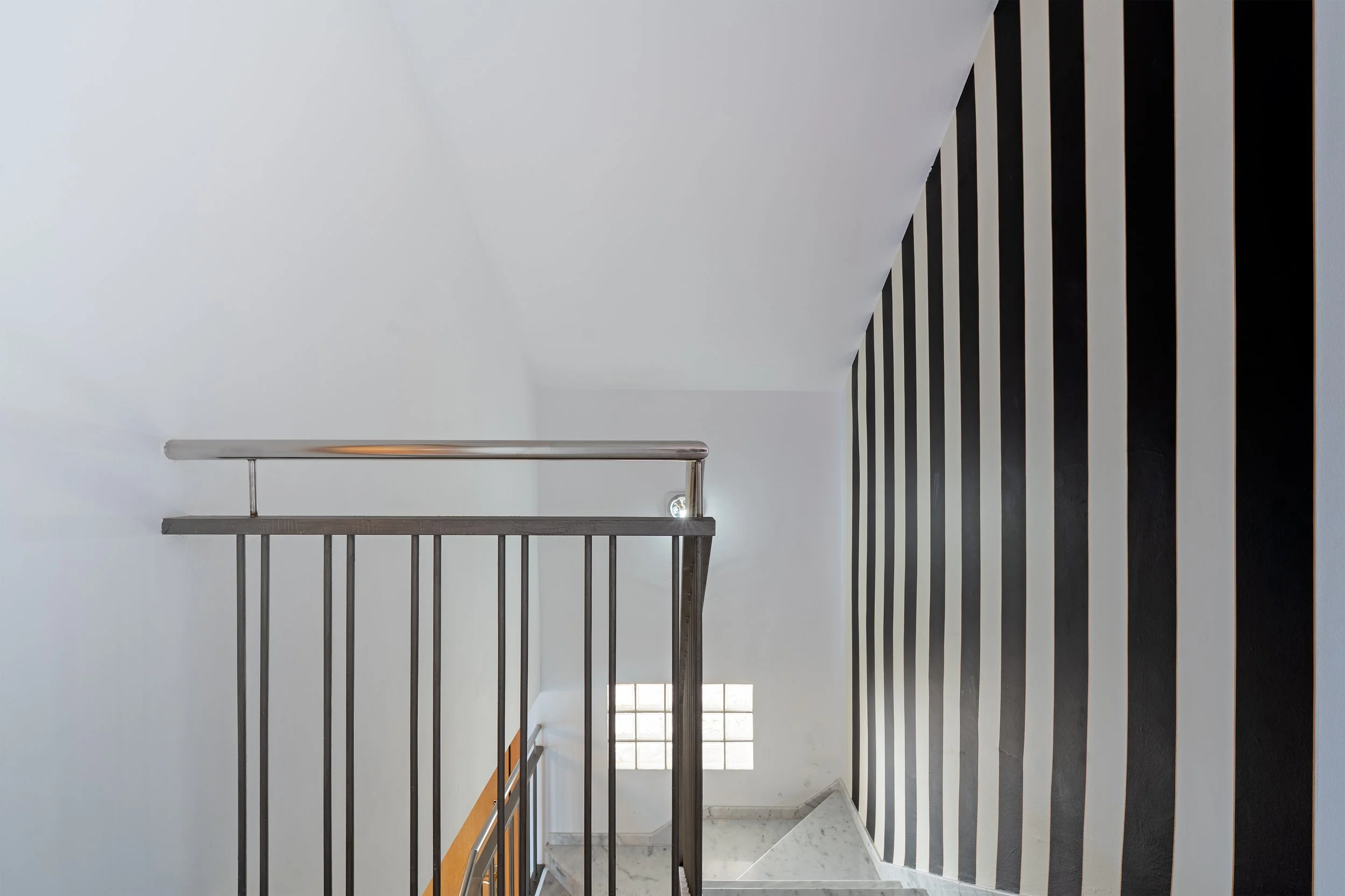 Interior view of a staircase with black and white striped wall on the right side, a white wall on the left, a stainless steel railing, and a small window at the bottom of the stairs.