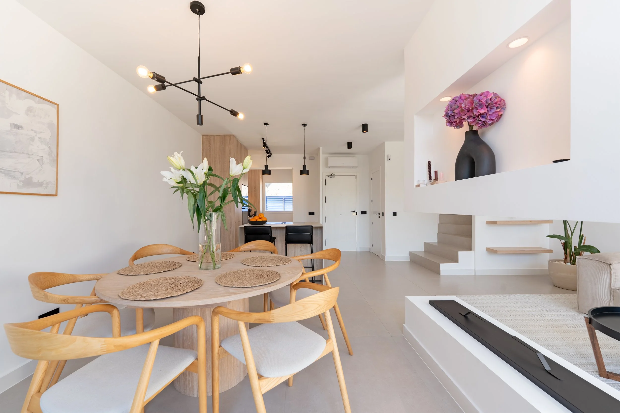 Open-concept living and dining area with white walls, wooden furniture, and black accents. A dining table with six chairs and a glass vase with white flowers is in the foreground. In the background, there is a kitchen with a small breakfast bar, blac