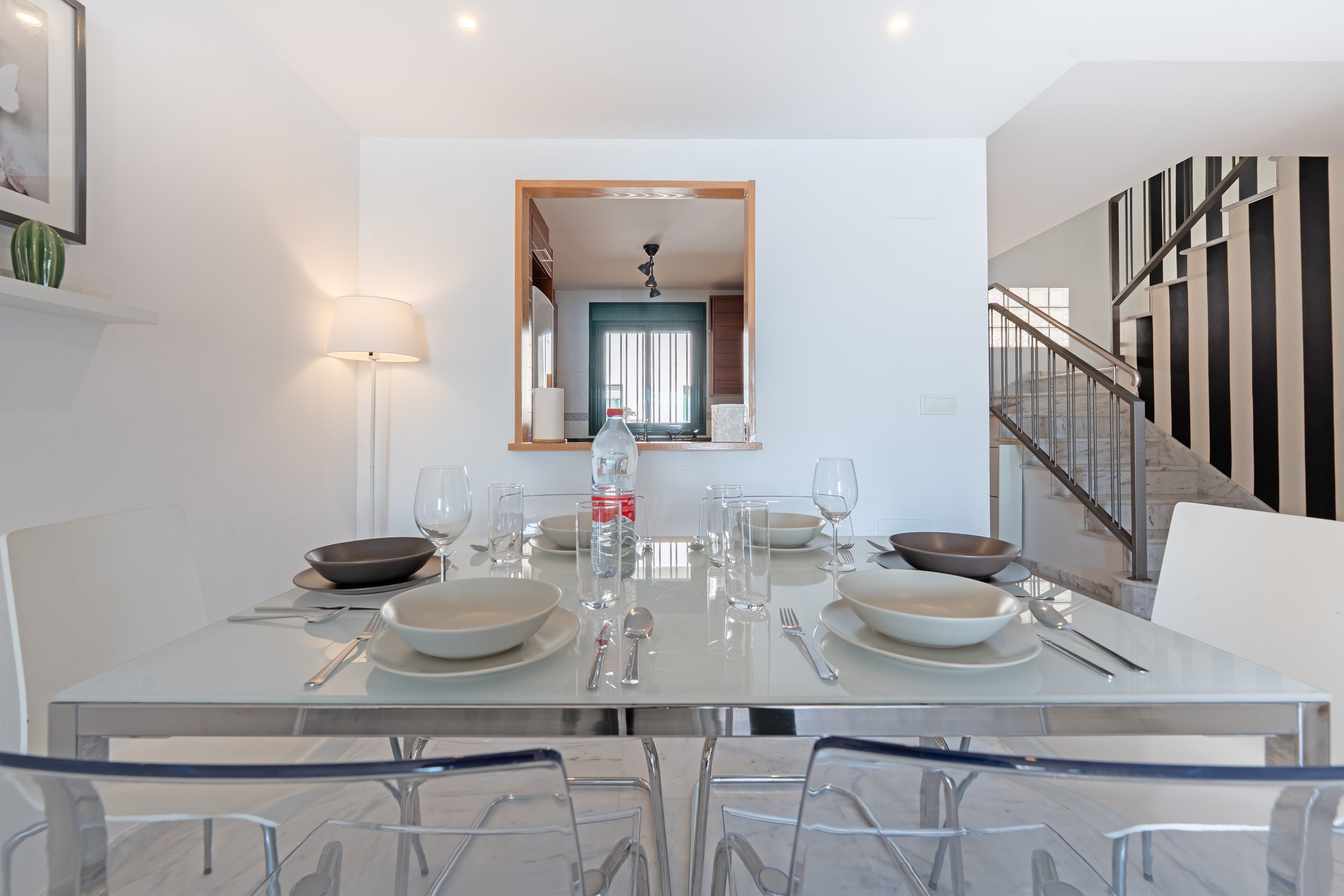Modern dining room with a white table set with bowls, plates, glasses, and silverware, and a backdrop of a kitchen visible through a window cutout, with a staircase to the right leading upstairs.