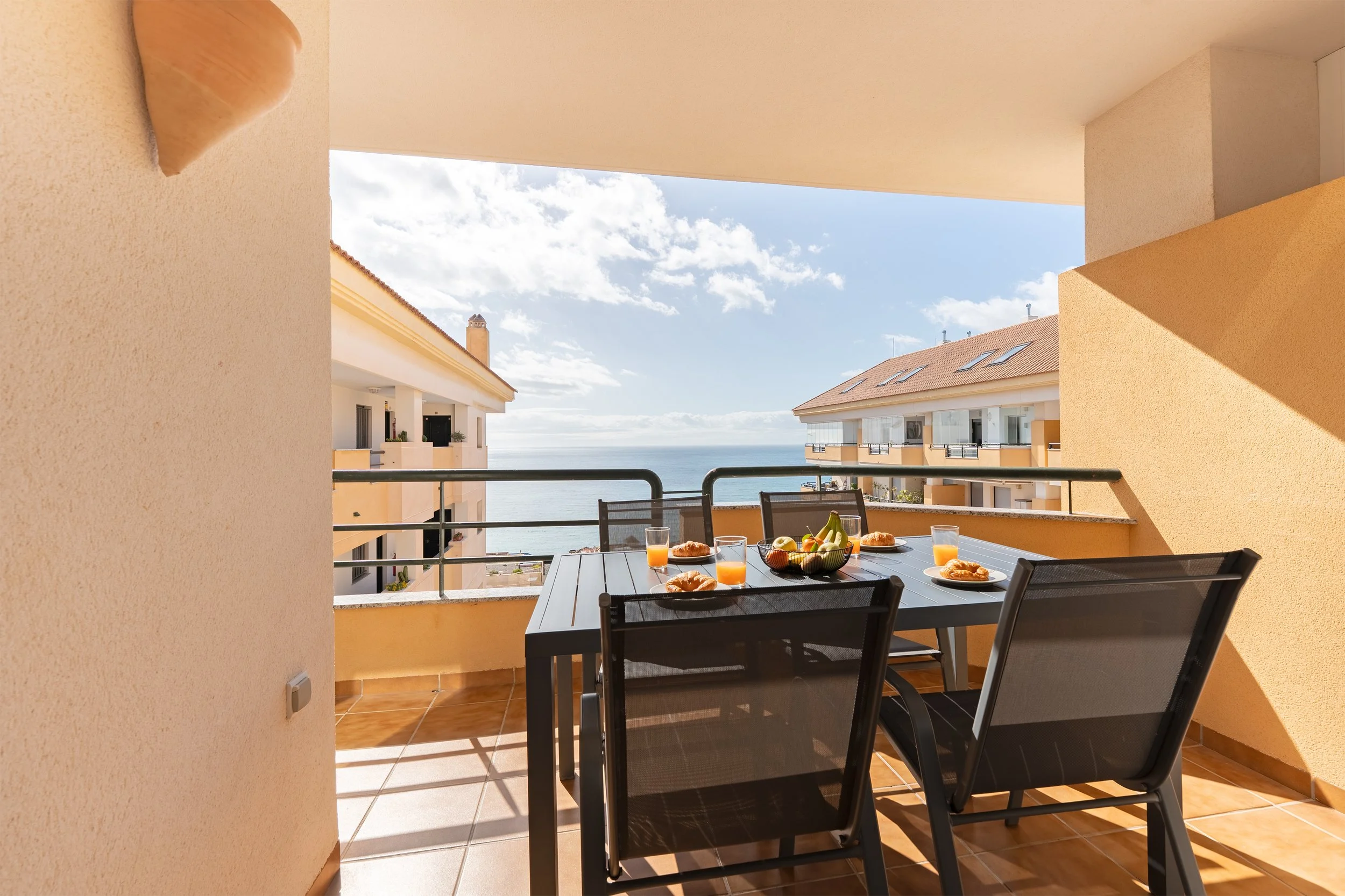 Balcony with a table set for breakfast, featuring plates with croissants, glasses of orange juice, a bowl of fruit including bananas and apples, overlooking neighboring apartment buildings and the ocean under a partly cloudy sky.