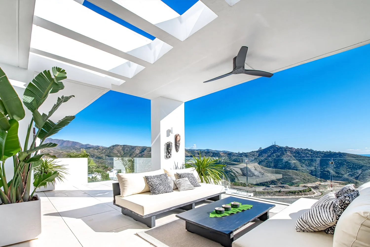 Modern outdoor patio with white furniture and cushions, large potted plants, glass railing, mountainous landscape, and blue sky.