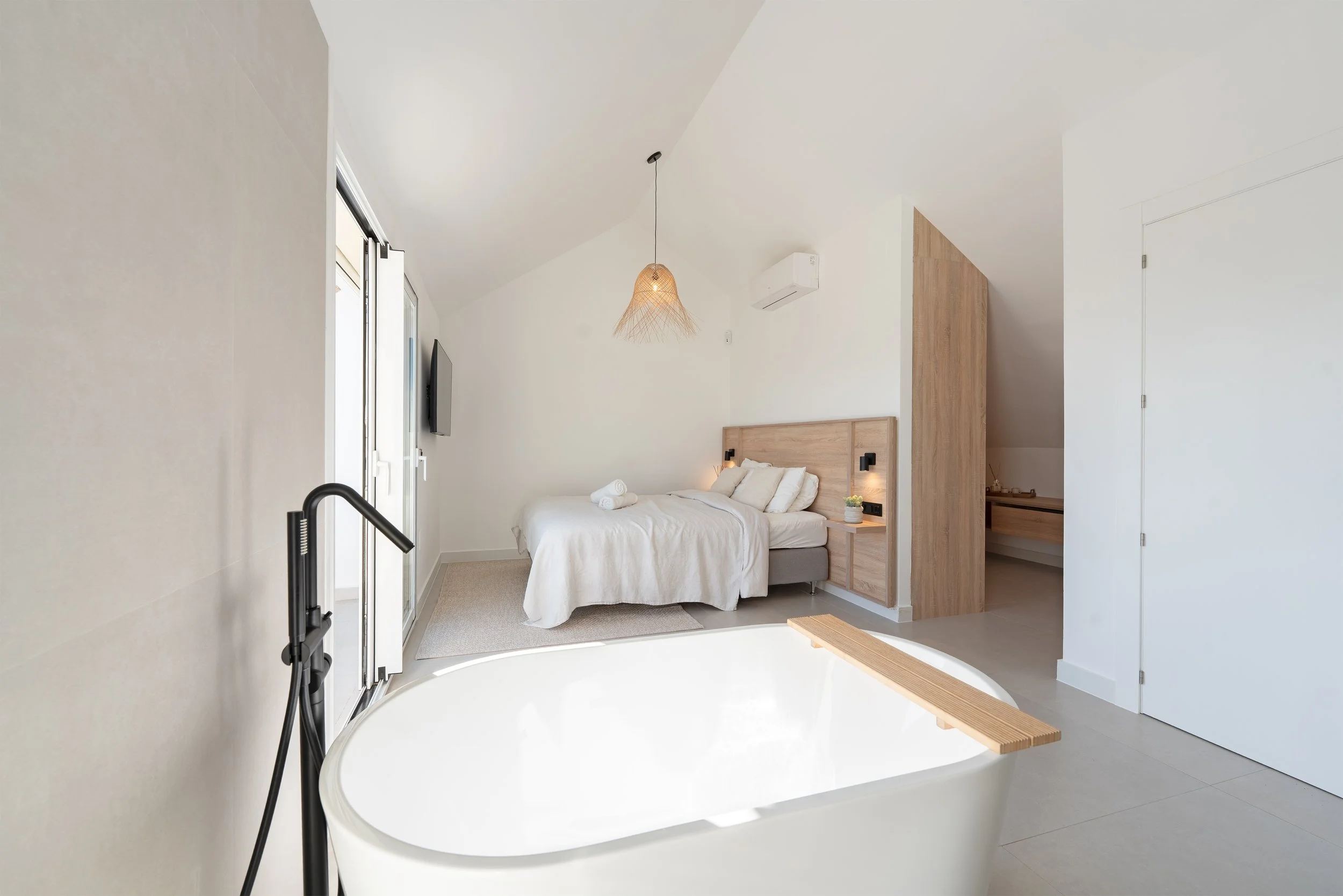 Modern hotel room with a white bed, wooden headboard, and a large bathtub in the foreground. The room features minimal decor, a pendant light, and a flat-screen TV on the wall.