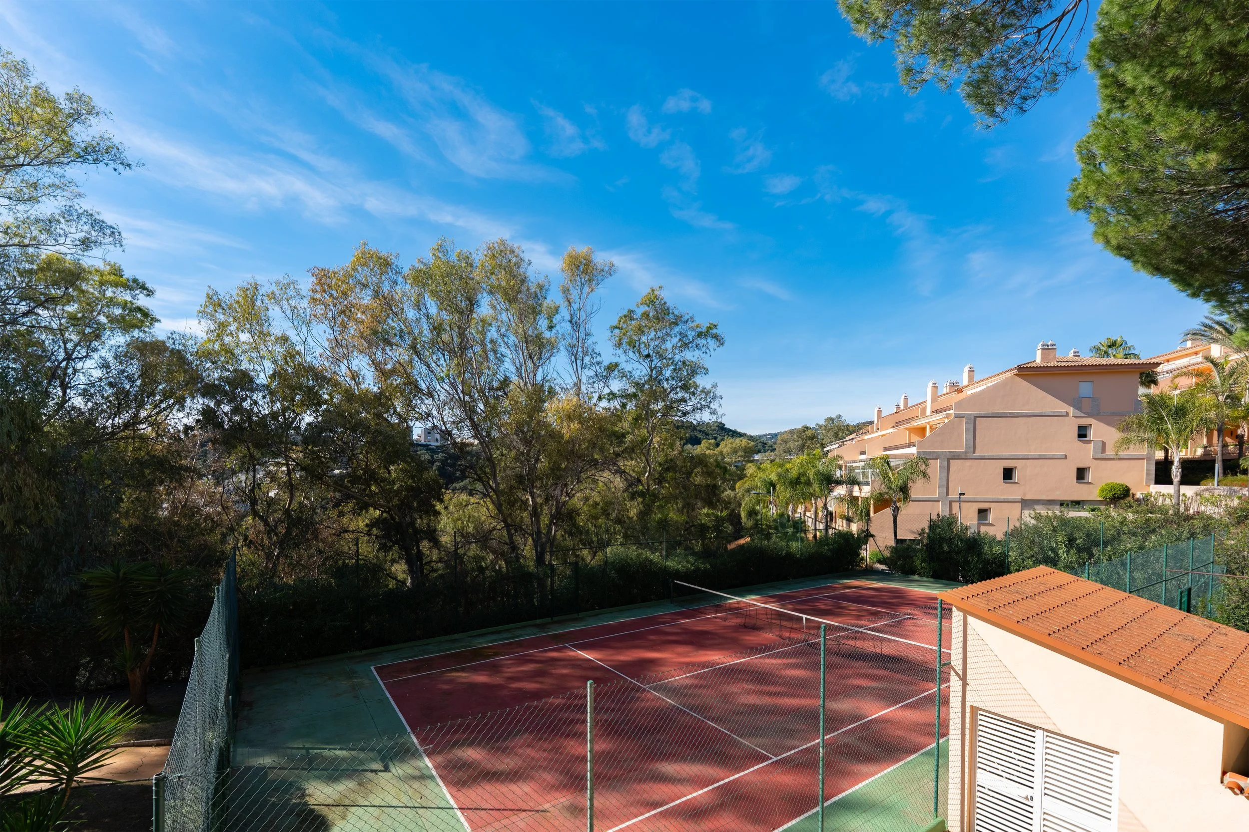 Empty tennis court with red and green surface, surrounded by a chain-link fence, situated near residential buildings with tan walls and red-tiled roofs, surrounded by trees and greenery under a blue sky with some clouds.