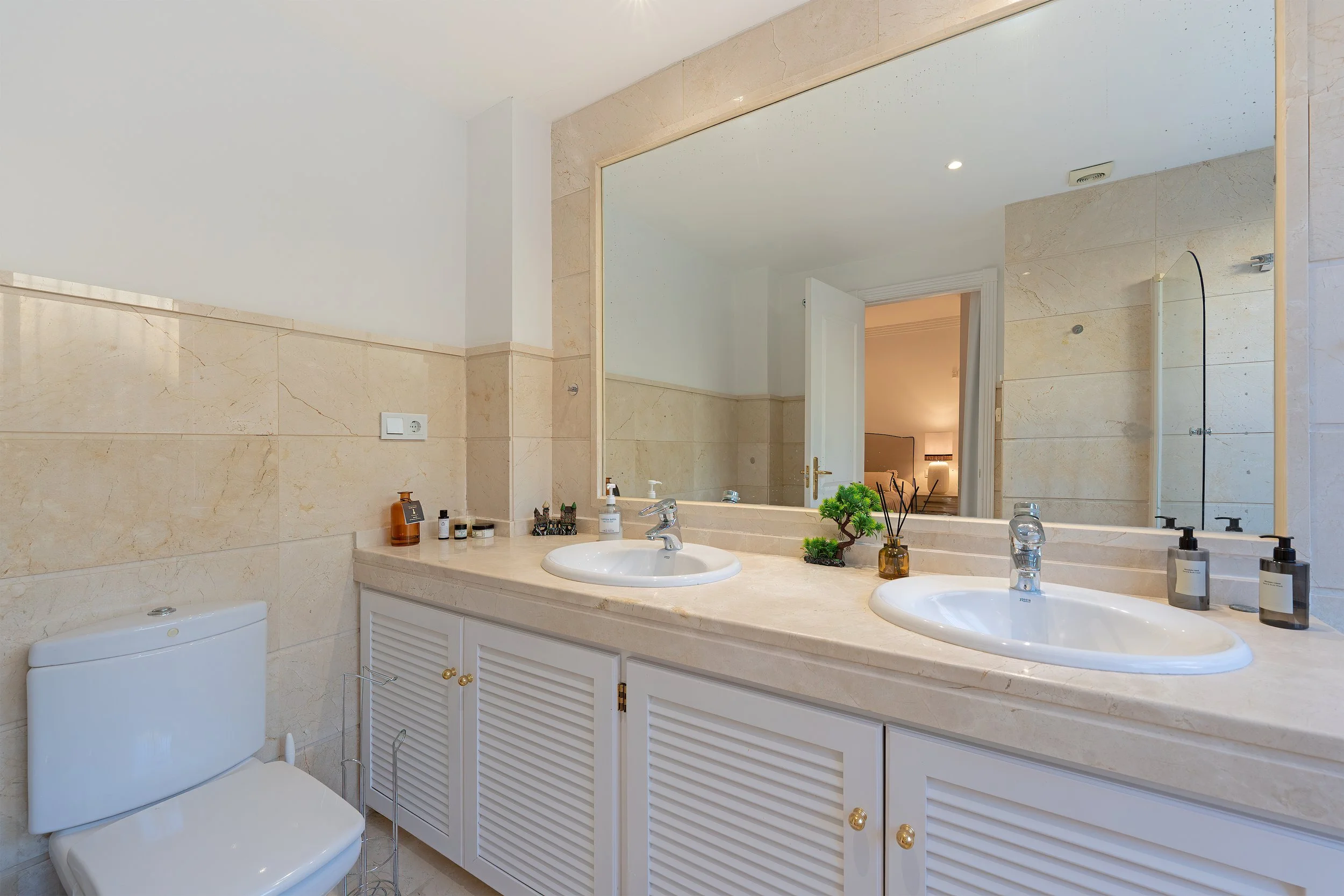 Bathroom with beige tiled walls, large mirror, twin sinks with soap and lotion bottles, small decorative plants, and a white toilet.