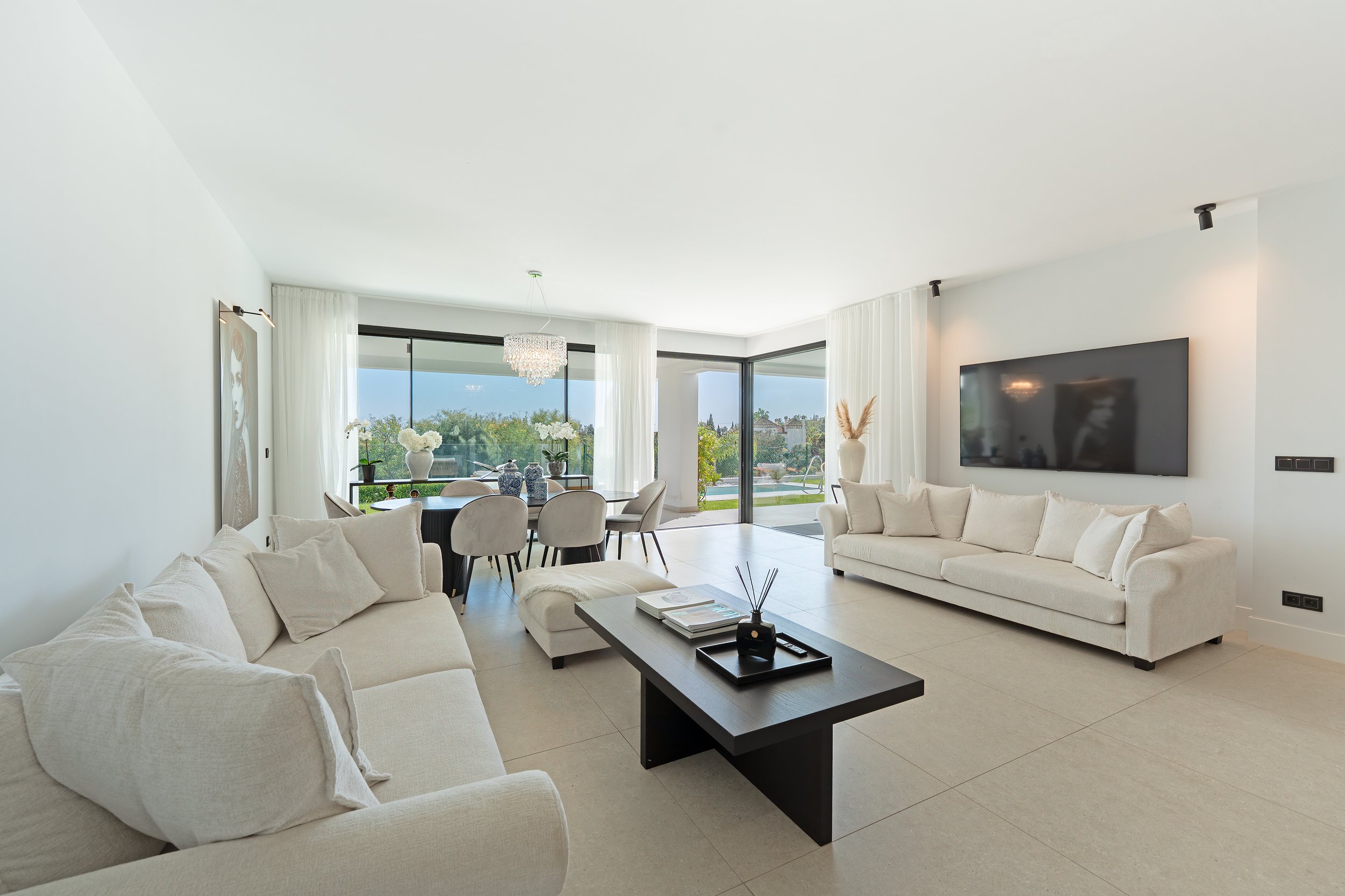 Bright living room with white sofas, a black coffee table, a dining area, large windows, white curtains, and a wall-mounted TV.