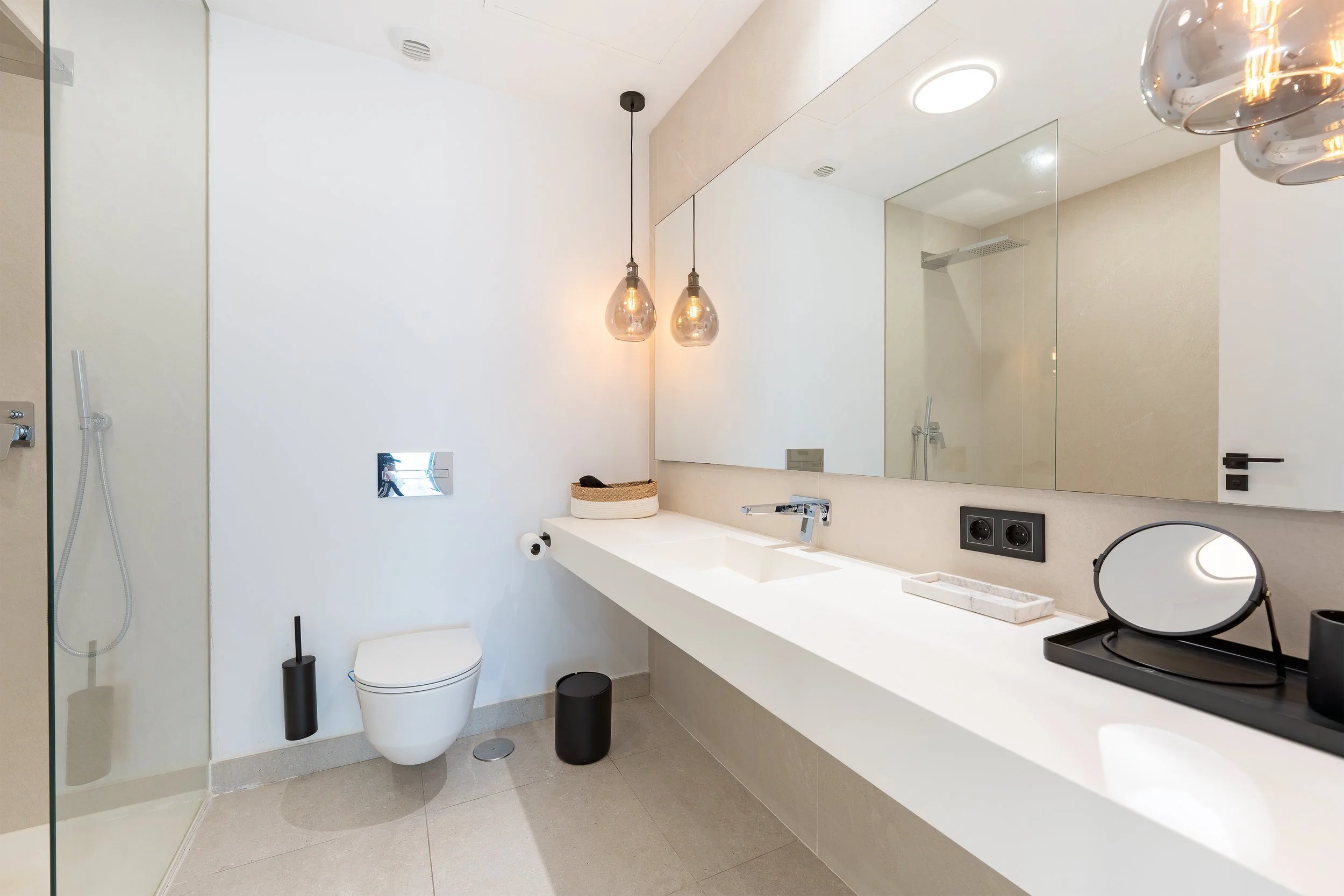 Modern minimalist bathroom with a wall-mounted toilet, a large mirror, a white countertop with a built-in sink, decorative pendant lights, and a glass shower enclosure.