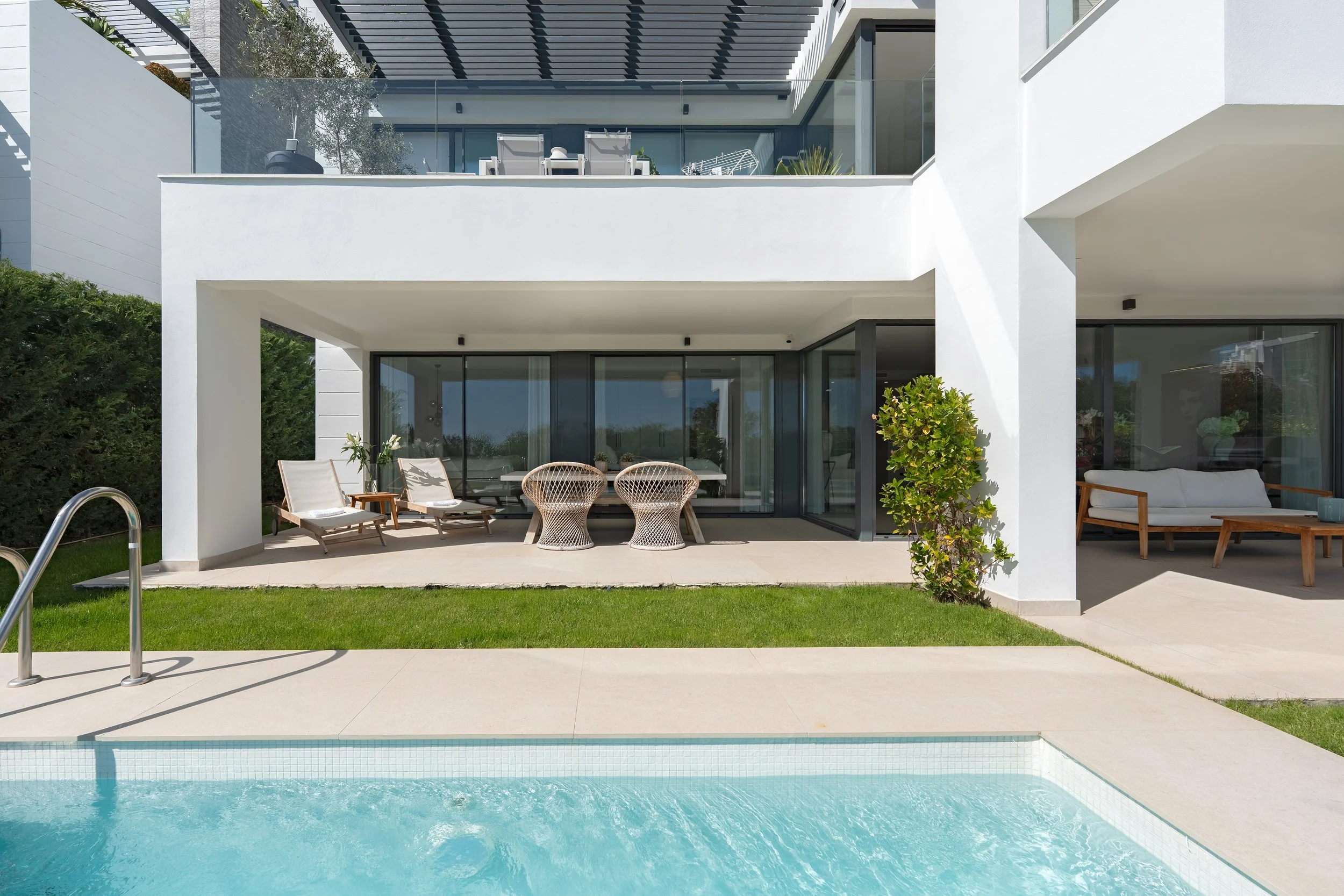 Modern two-story house with a backyard pool and patio area. There are lounge chairs, wicker chairs, and a sofa on the patio. The house features glass sliding doors and balconies with outdoor furniture.