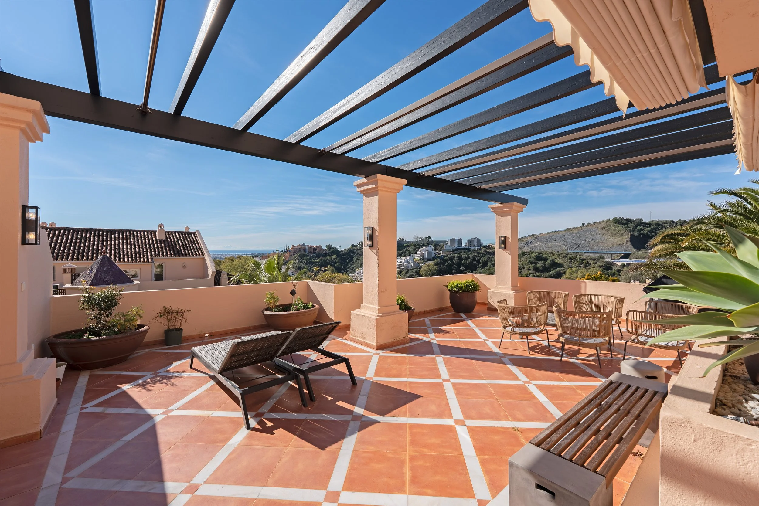 Sunlit outdoor terrace with terracotta tile floor, potted plants, and wicker patio furniture, overlooking a hilly landscape and blue sky.