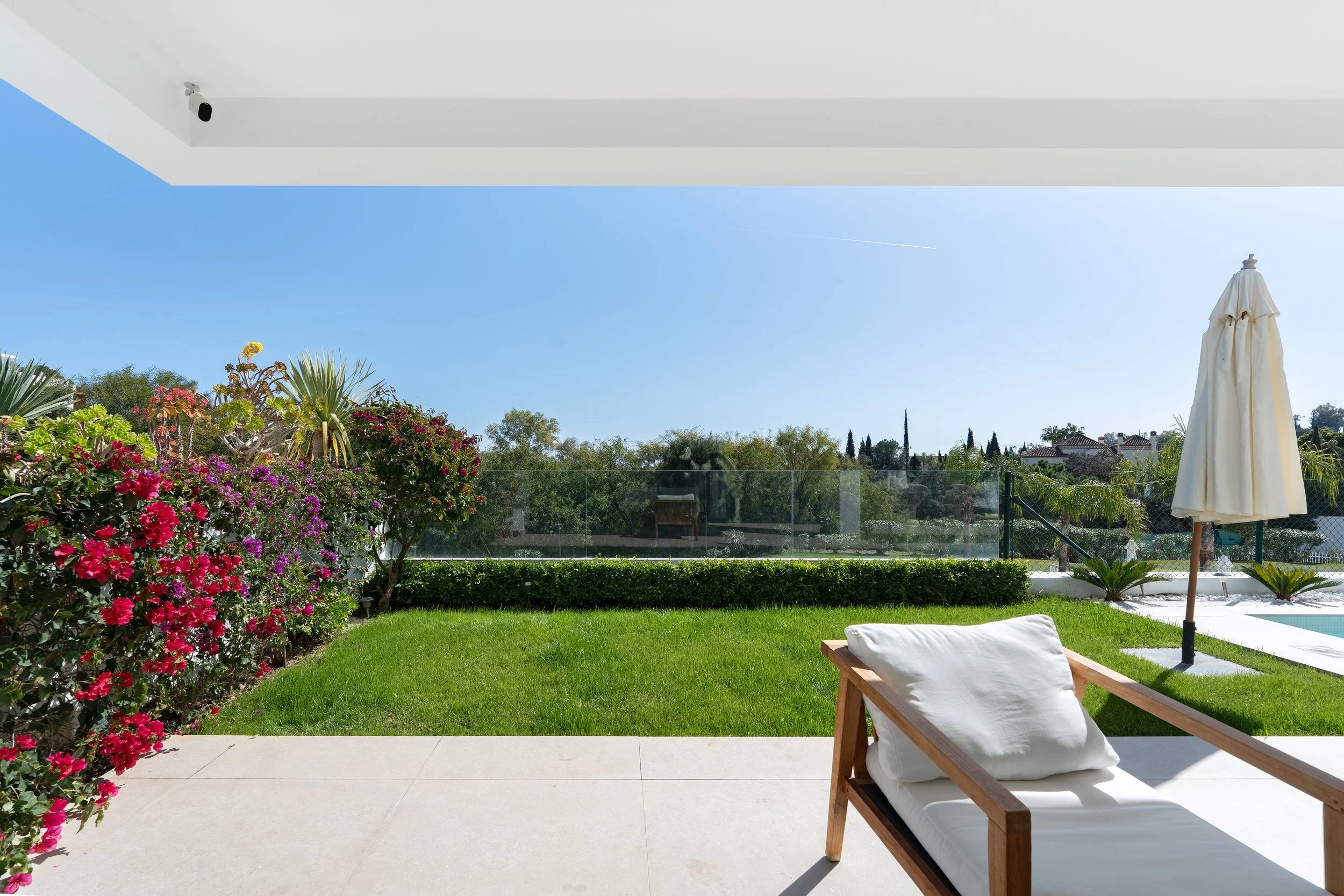 View of a backyard with a pool, lush green grass, vibrant flowering bushes, a wooden lounge chair with a white cushion, and a closed patio umbrella under a clear blue sky.