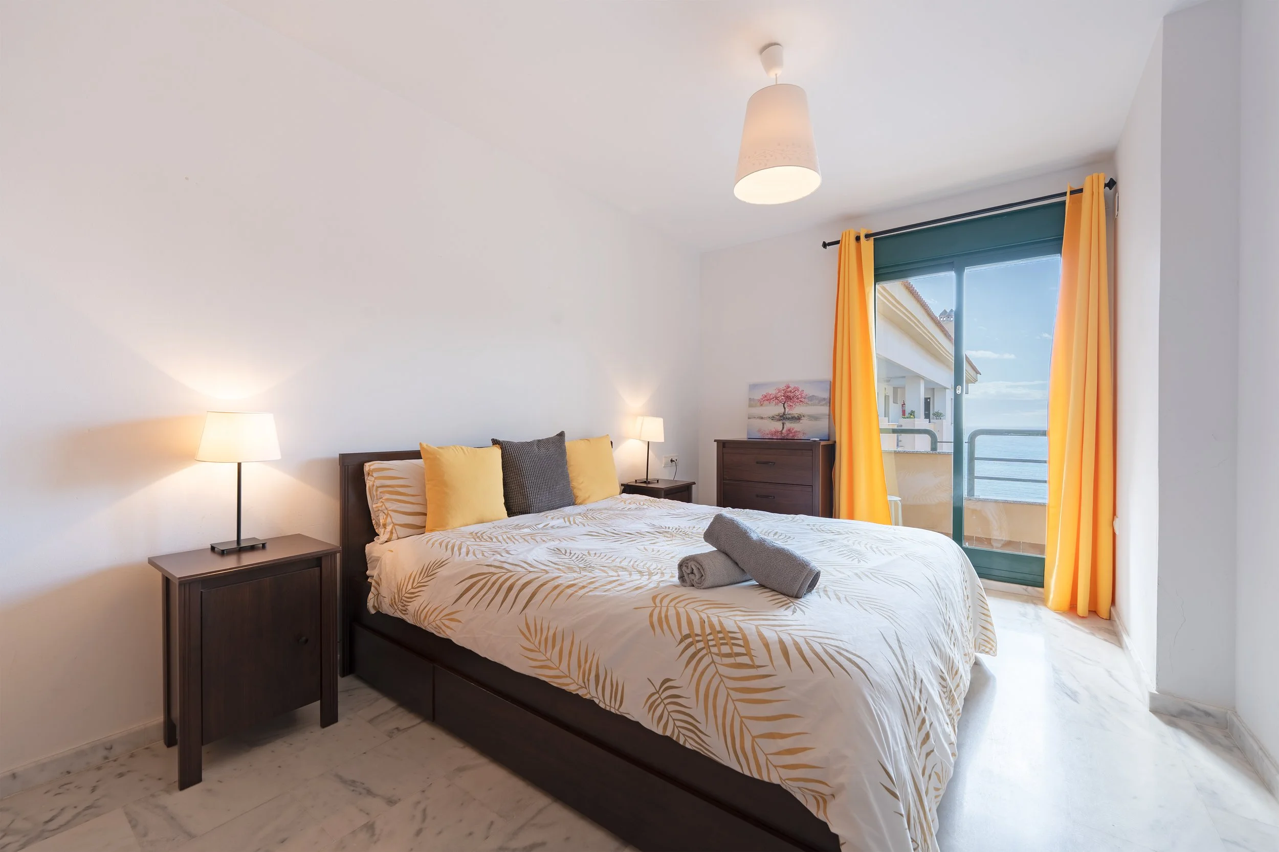 Bedroom with a bed, nightstand, lamps, dresser, and sliding glass door with yellow curtains leading to a balcony with ocean view.