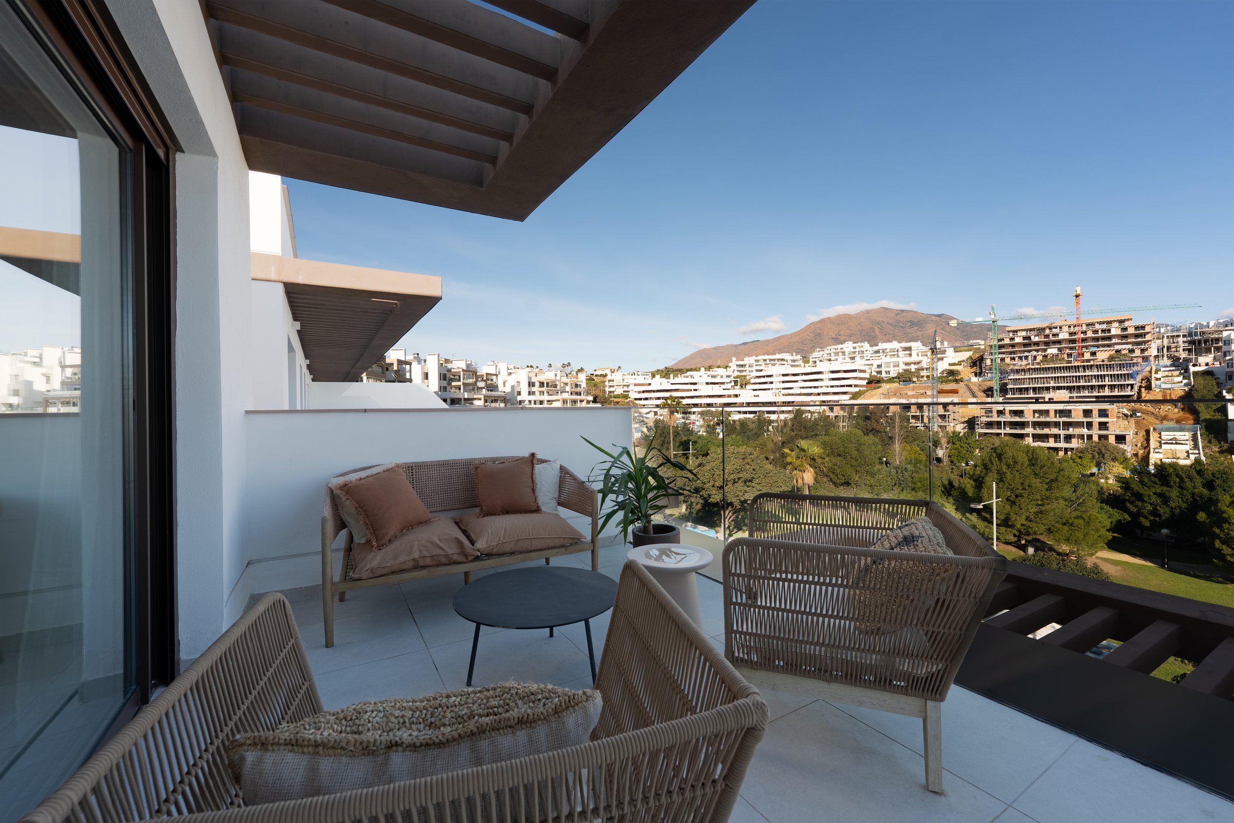 Balcony with outdoor furniture including a wicker sofa with cushions, a wicker chair, a small round table, a potted plant, and a scenic view of white apartment buildings, green trees, and mountains under a clear blue sky.