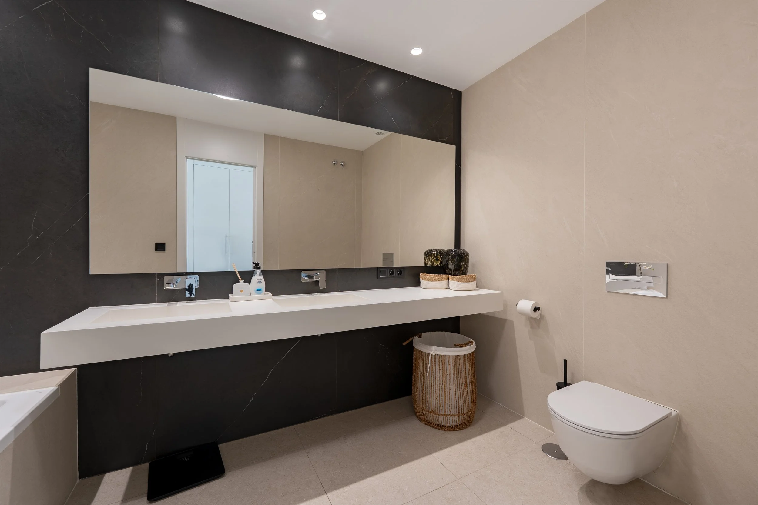 Modern bathroom with large mirror, white vanity, dual sinks, black and beige walls, decorative vases, laundry basket, toilet, and recessed ceiling lights.