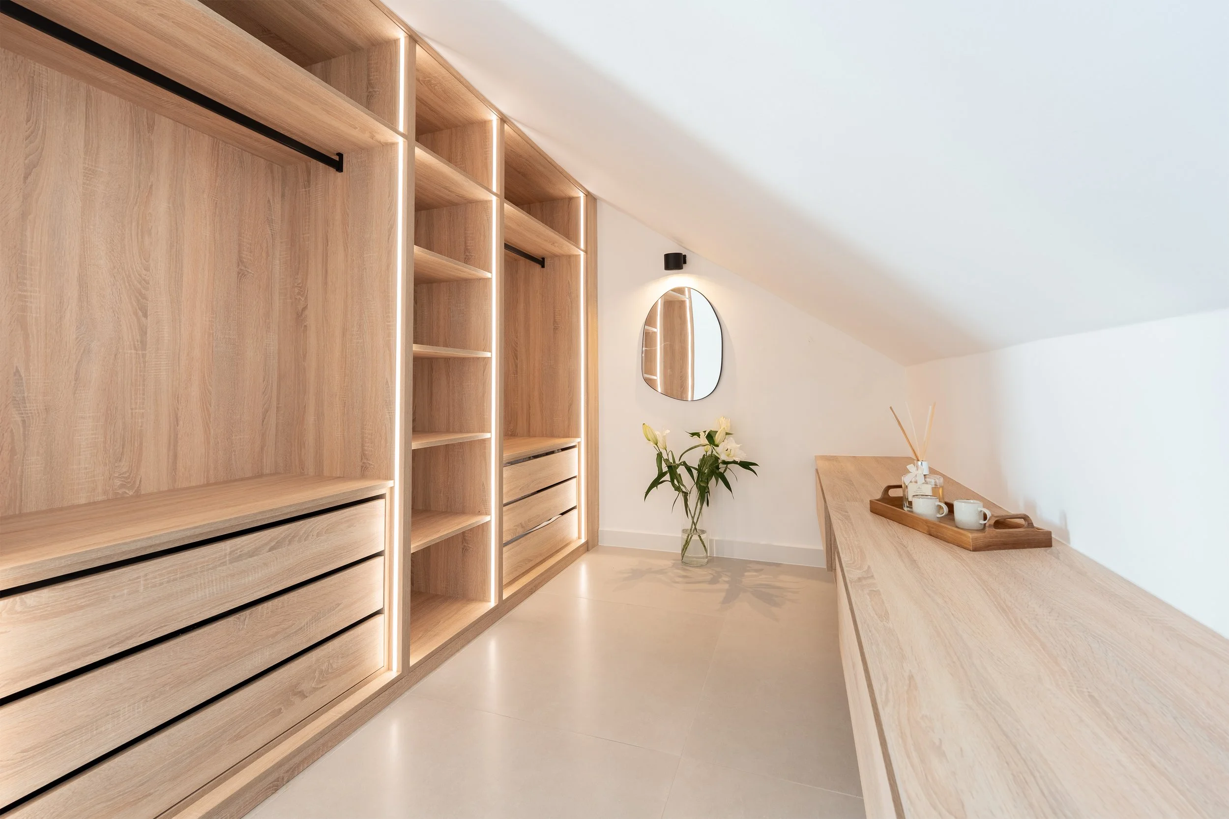 Minimalist walk-in closet with wooden shelves, drawers, and a long countertop. A vase with white flowers, a mirror, and a tray with cups and diffuser sticks on the countertop. Light-colored flooring and white walls.