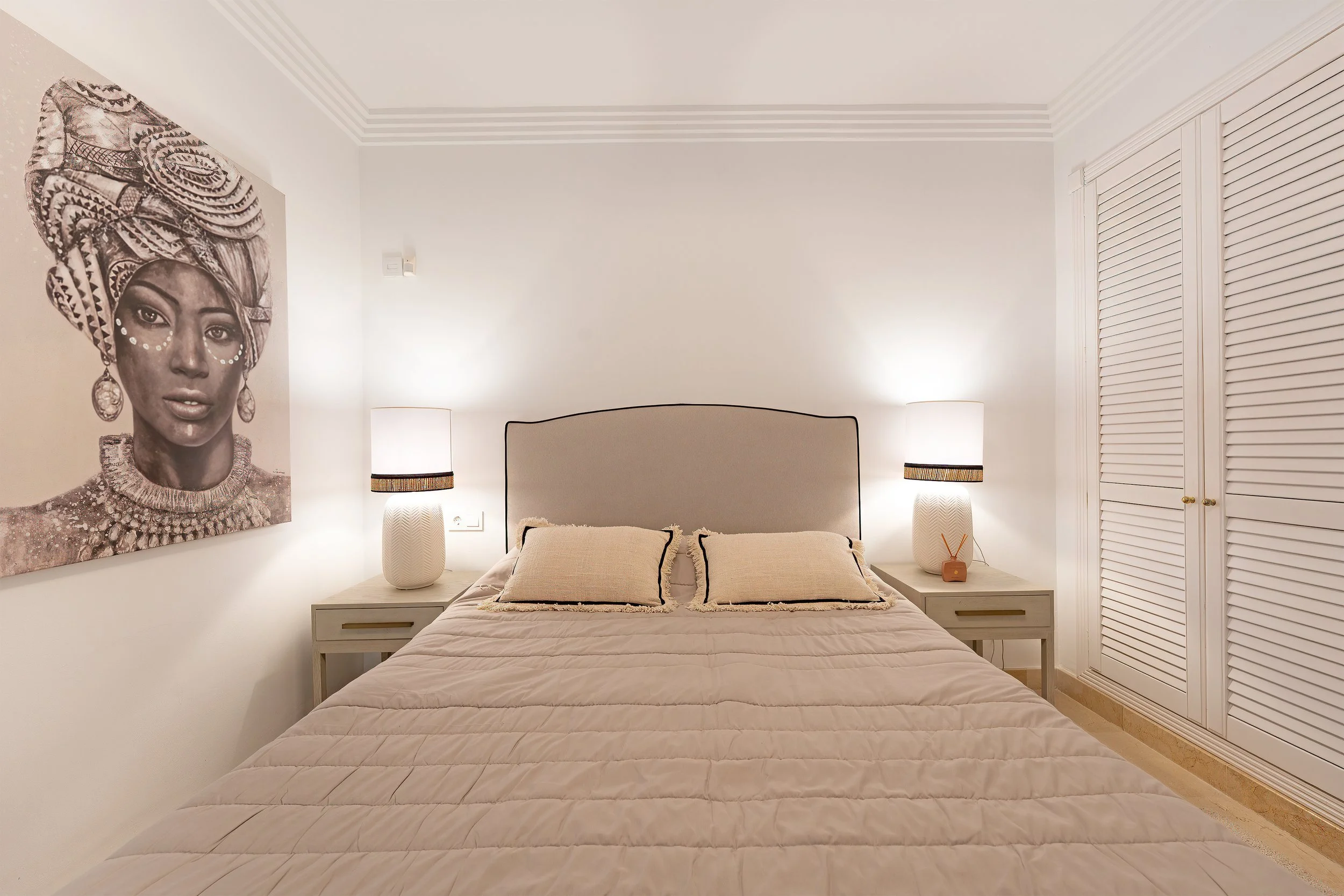 A bedroom with a bed, two pillows, two nightstands with lamps, a large painting of a woman wearing a headwrap and jewelry, white closet doors, and beige walls.