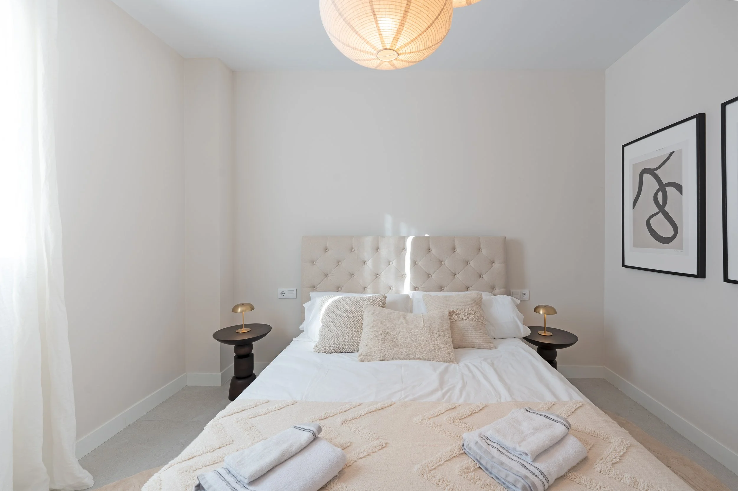 Modern bedroom with white walls, beige upholstered headboard, white bedding, decorative pillows, black nightstands with gold lamps, abstract black and white artwork, and neatly folded towels on the bed.