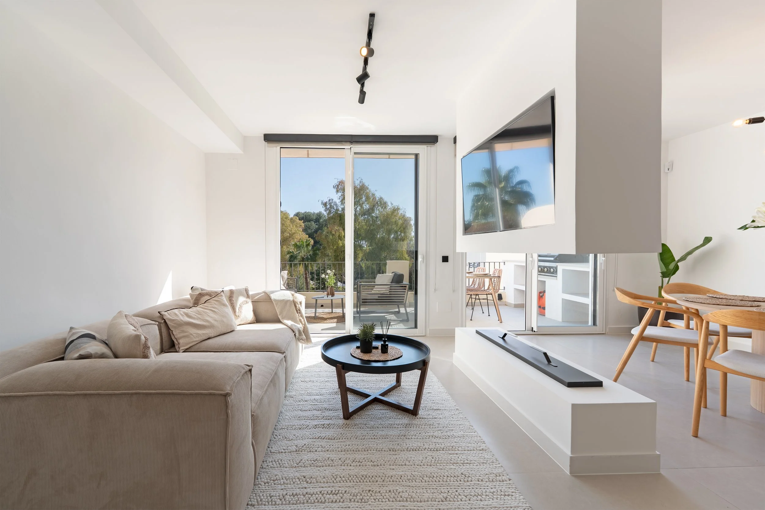 Modern living room with beige sofa, black round coffee table, white textured rug, large glass sliding doors opening to a balcony with outdoor seating and scenic greenery, mounted flat-screen TV, white walls, and minimalistic decor.