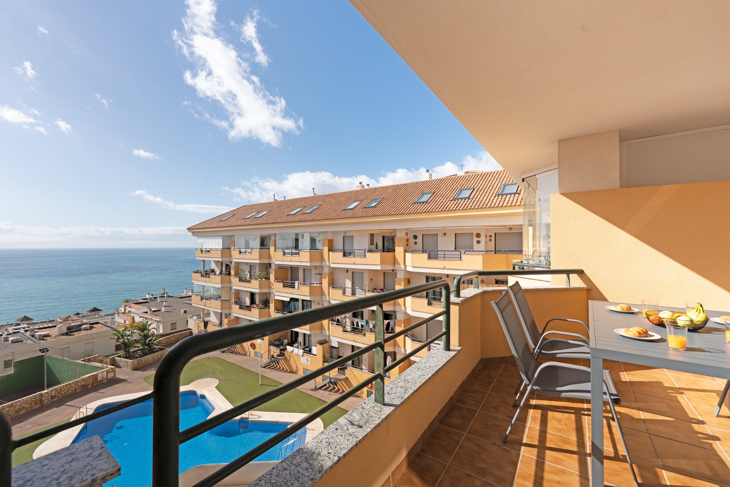 Balcony with table and chairs overlooking a hotel complex and ocean on a sunny day.
