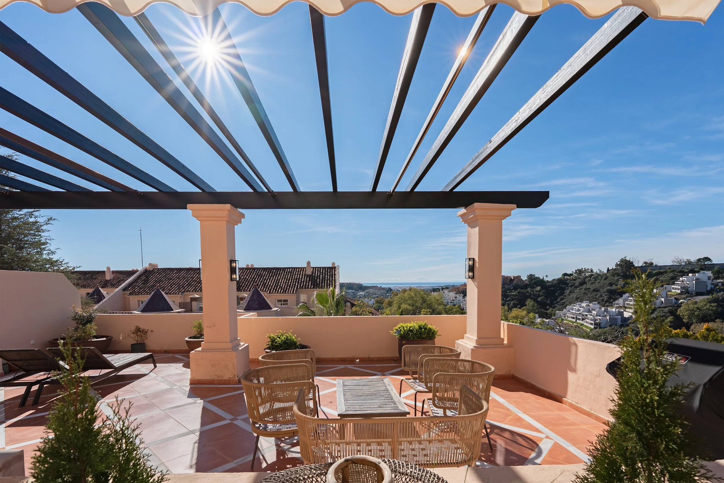 Outdoor rooftop patio with a table and six chairs, potted plants, a barbecue grill, and a view of houses and green hills under a bright blue sky.