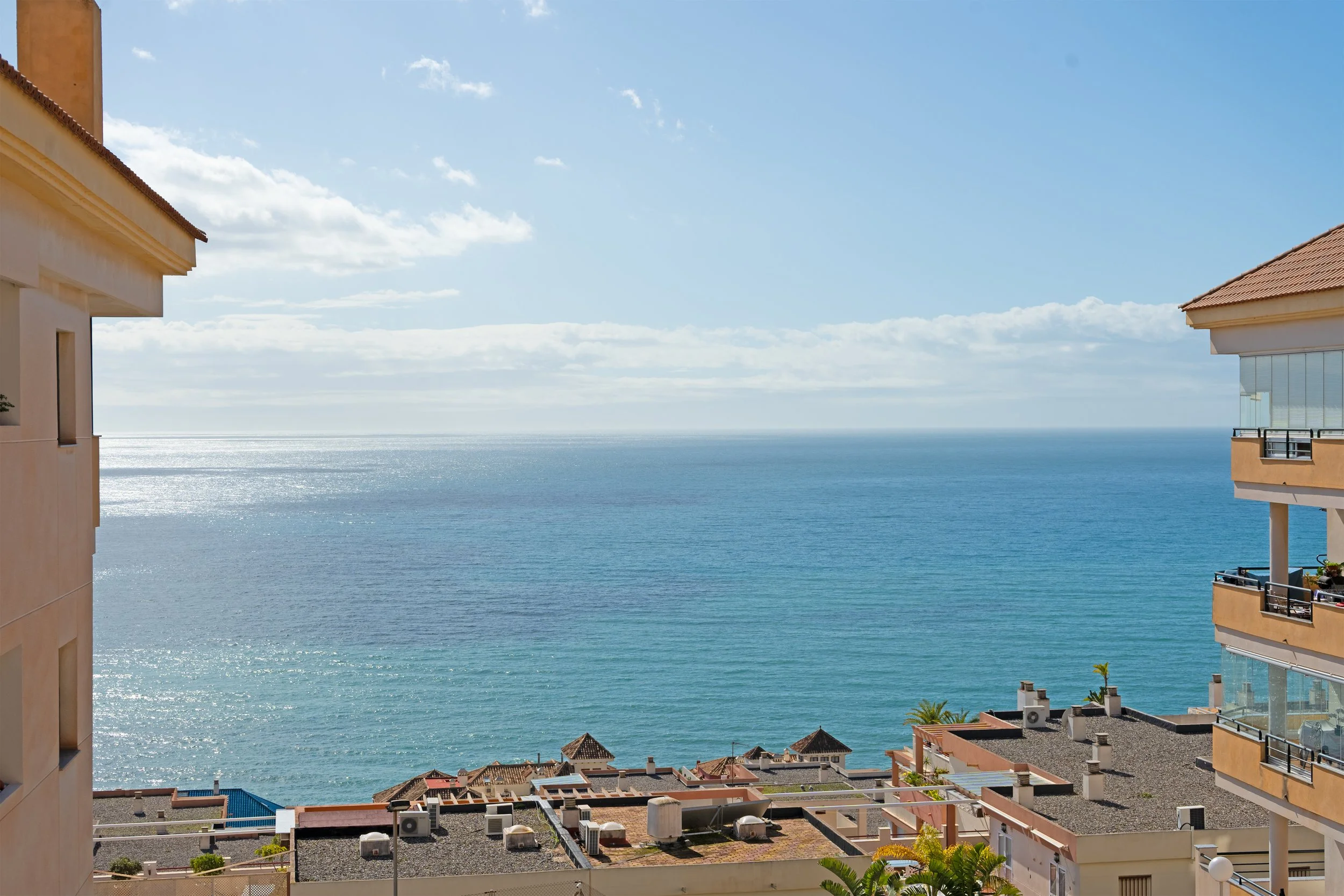 View of the ocean with a partly cloudy sky, seen from a balcony between two buildings with tan and orange exteriors.