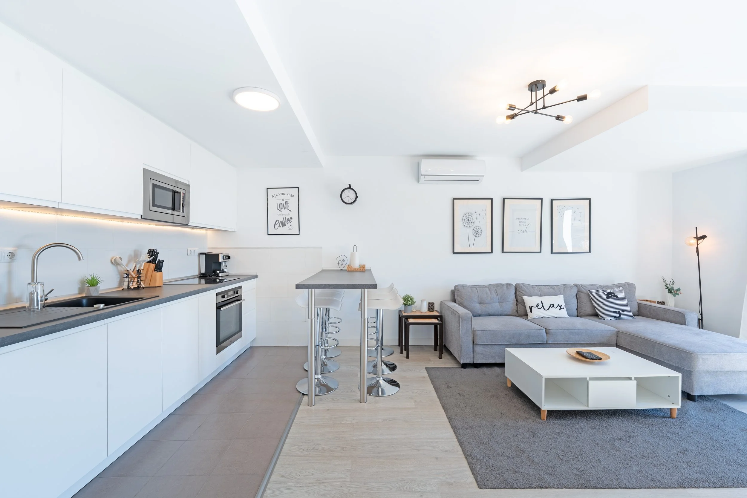 Modern living room and kitchen with white cabinets, gray couch, coffee table, artwork, and bar stools.