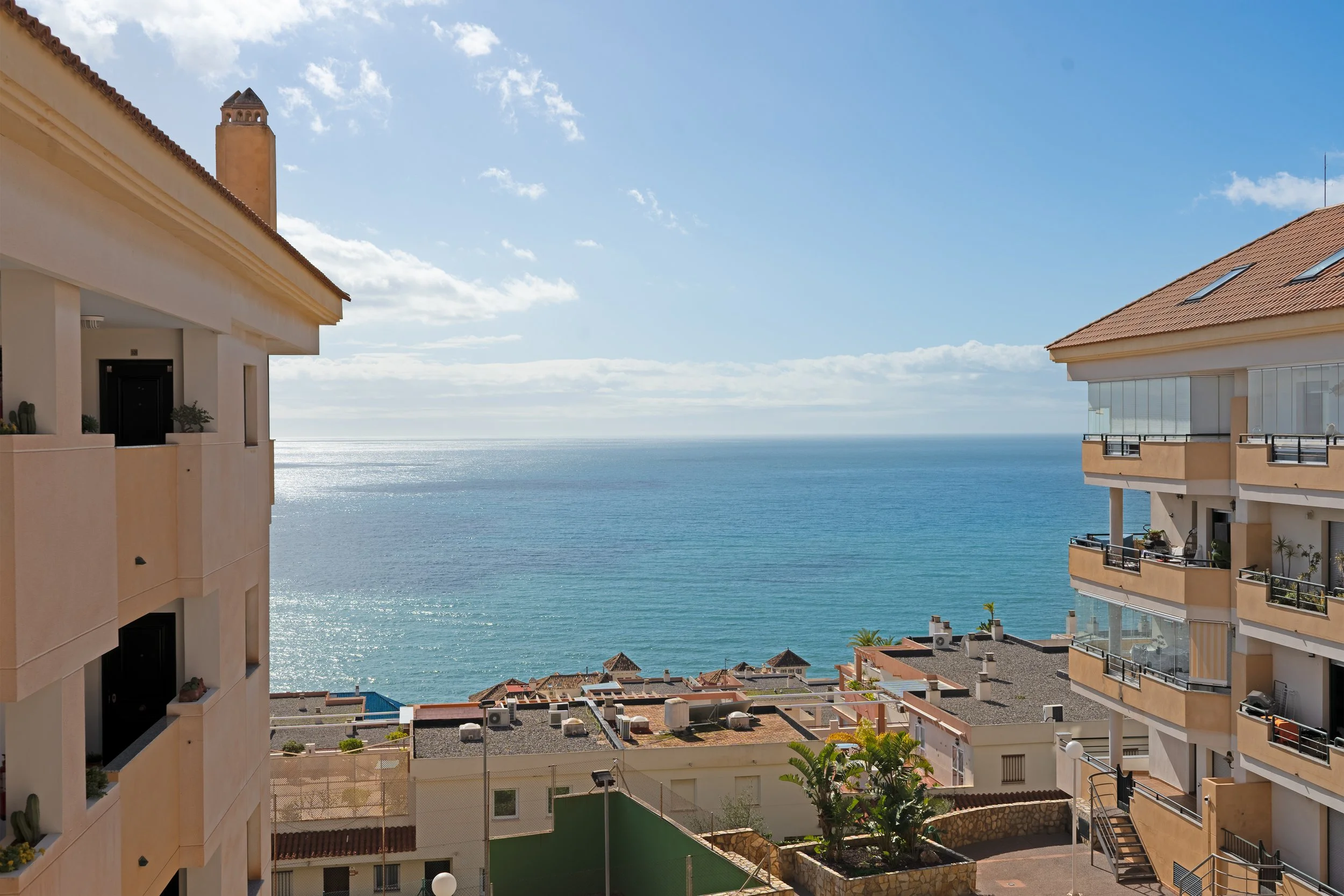 View of the ocean from a balcony between two beige residential buildings with balconies, some with plants, in a sunny setting with a partly cloudy sky.