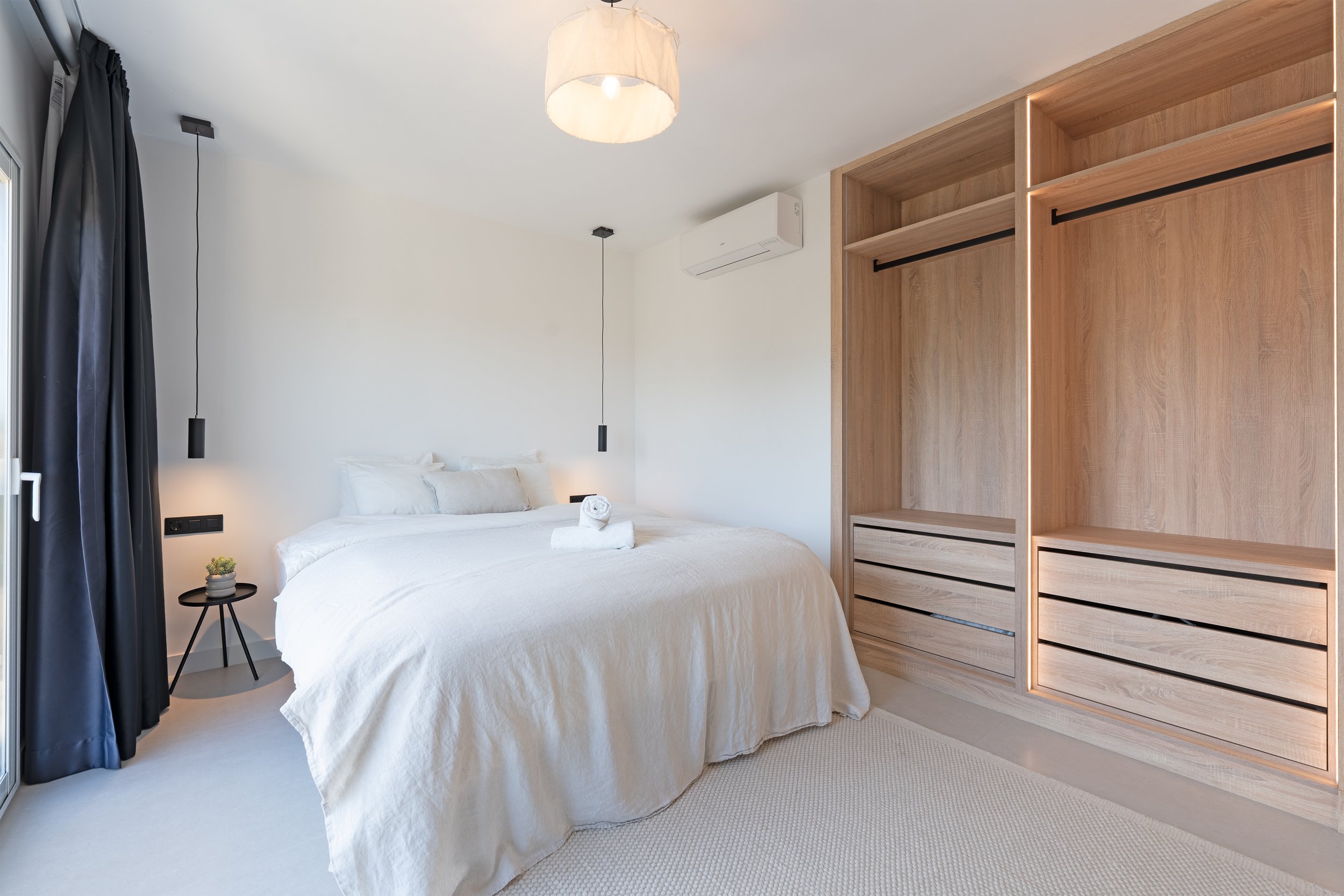 A modern, minimalist bedroom with a white bed, black curtains, wooden closet, and black pendant lights.
