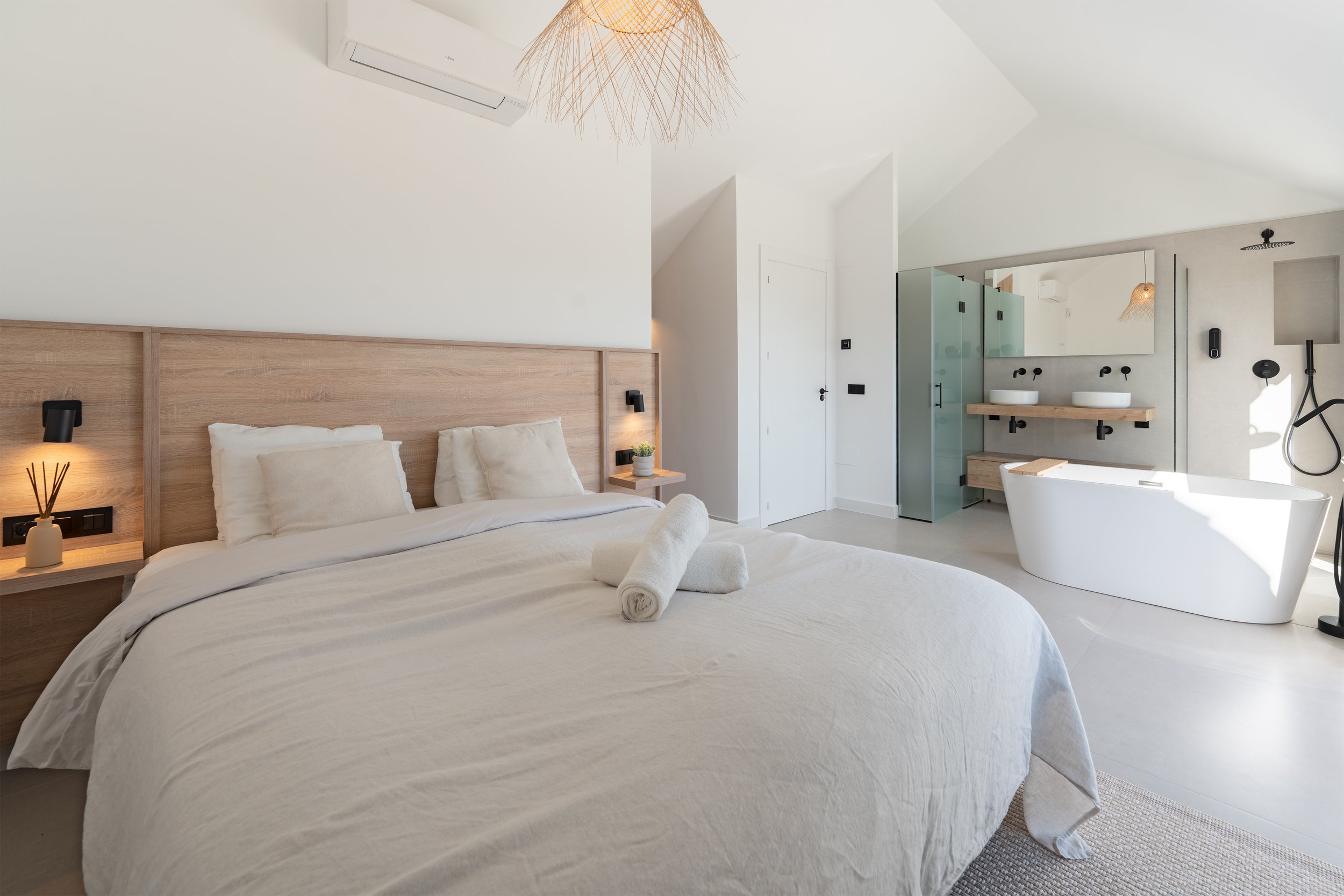 Modern bedroom with a large bed featuring white linens and pillows, wooden headboard, two rolled towels on the bed, hanging light fixture, bathroom area with two sinks and bathtub, glass shower enclosure, and minimalist decor.