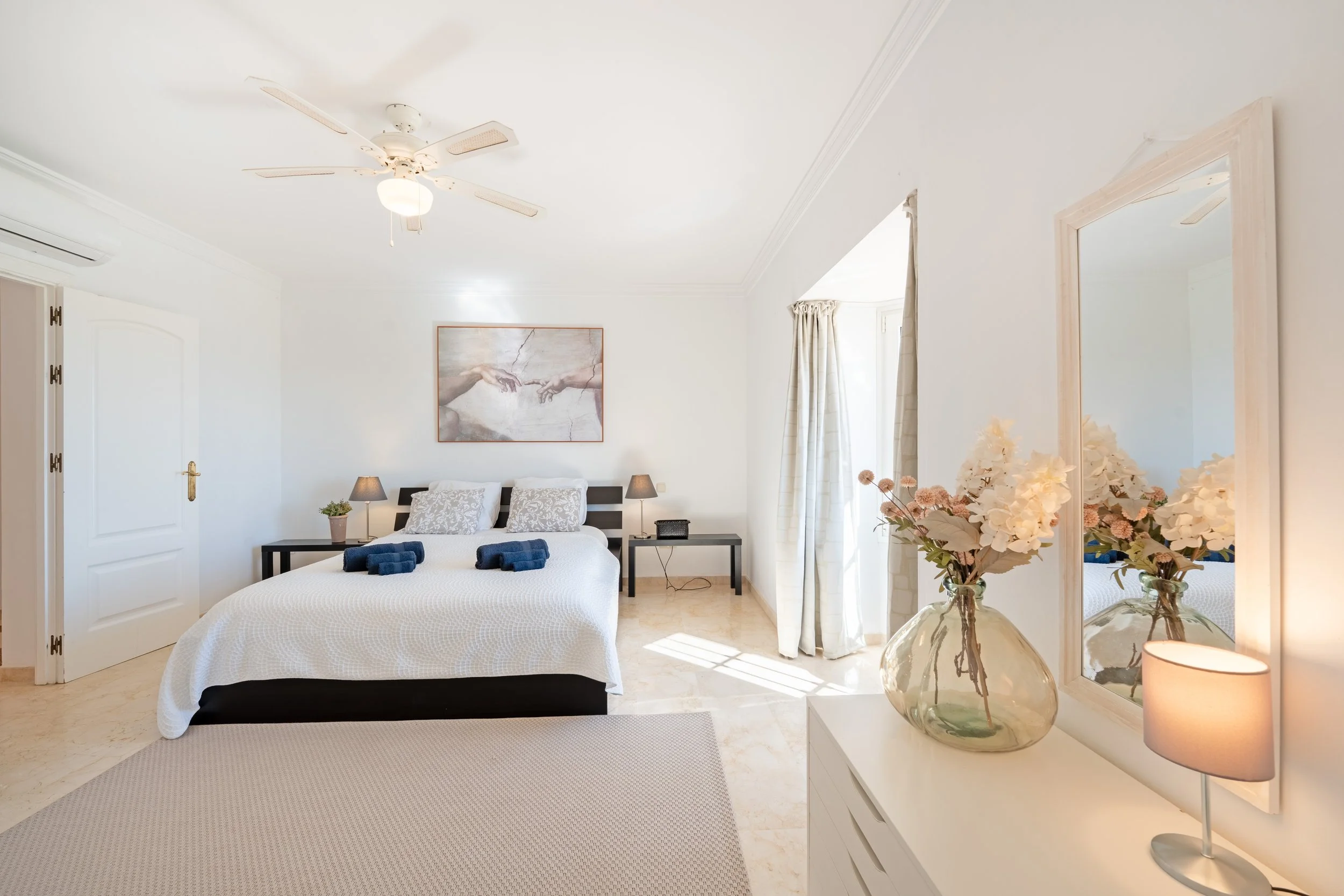 Bright hotel bedroom with white walls, a bed with white bedding and two pillows, wall art above the bed, bedside tables with lamps, a vase with flowers, a mirror, and a source of natural light from a window with curtains.