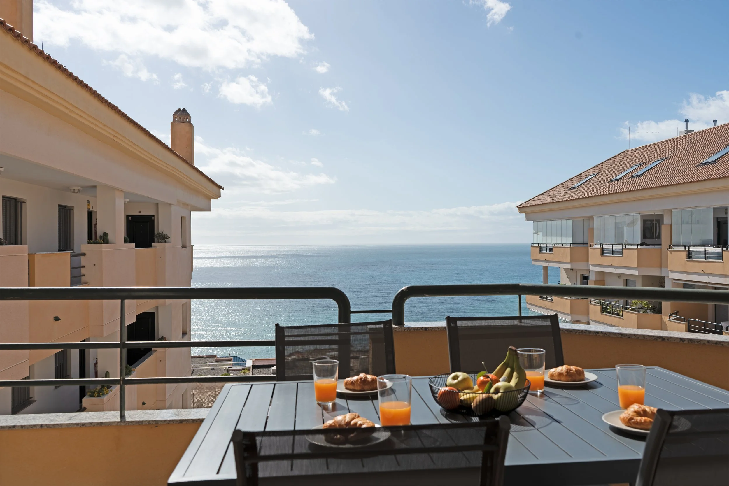 Balcony with a table set for breakfast, including glasses of orange juice, plates of pastries, and a fruit bowl with bananas and apples, overlooking the ocean with modern buildings on either side.