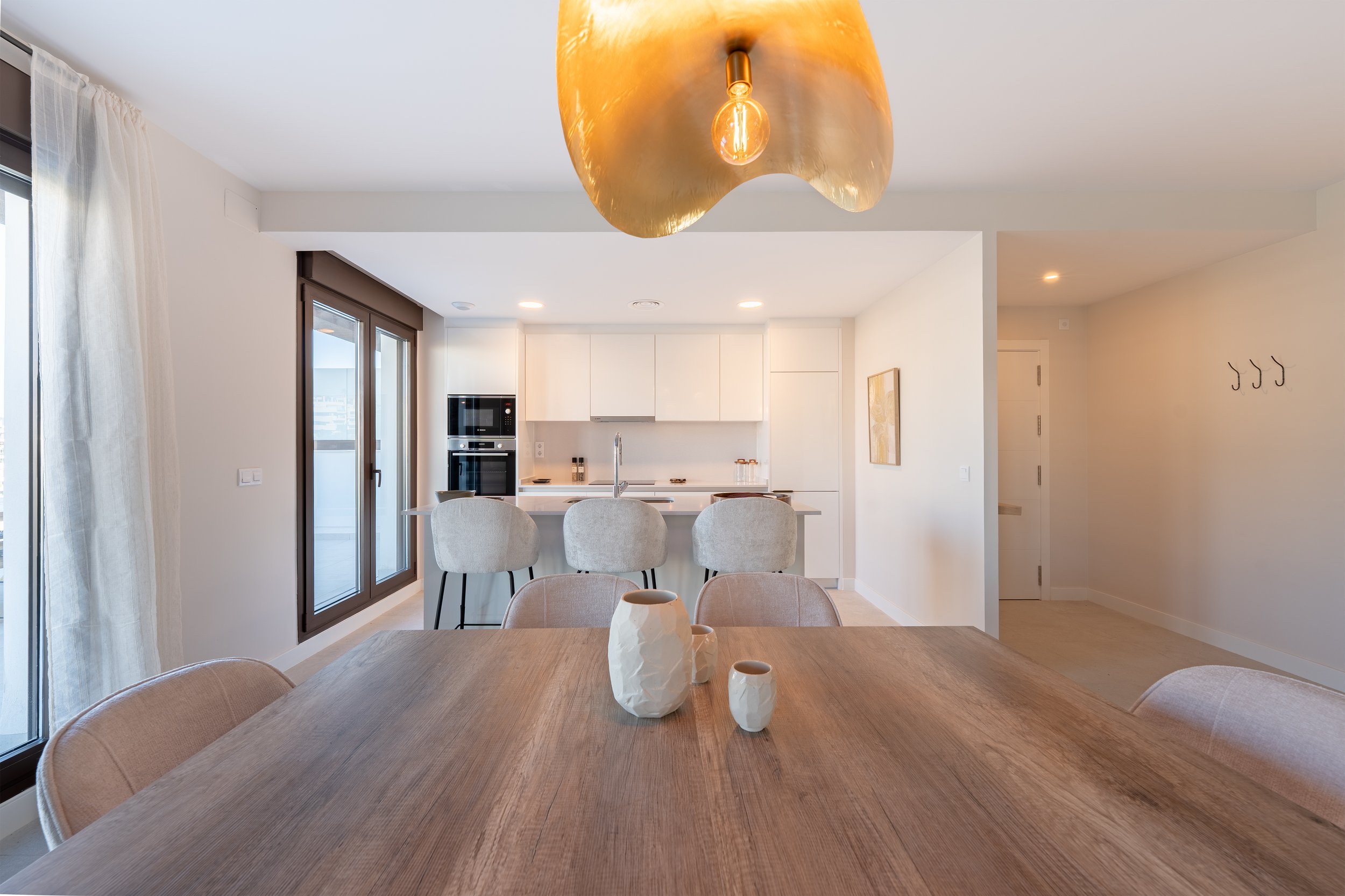 Modern open-plan dining and kitchen area with a wooden dining table, white chairs, and a gold pendant light