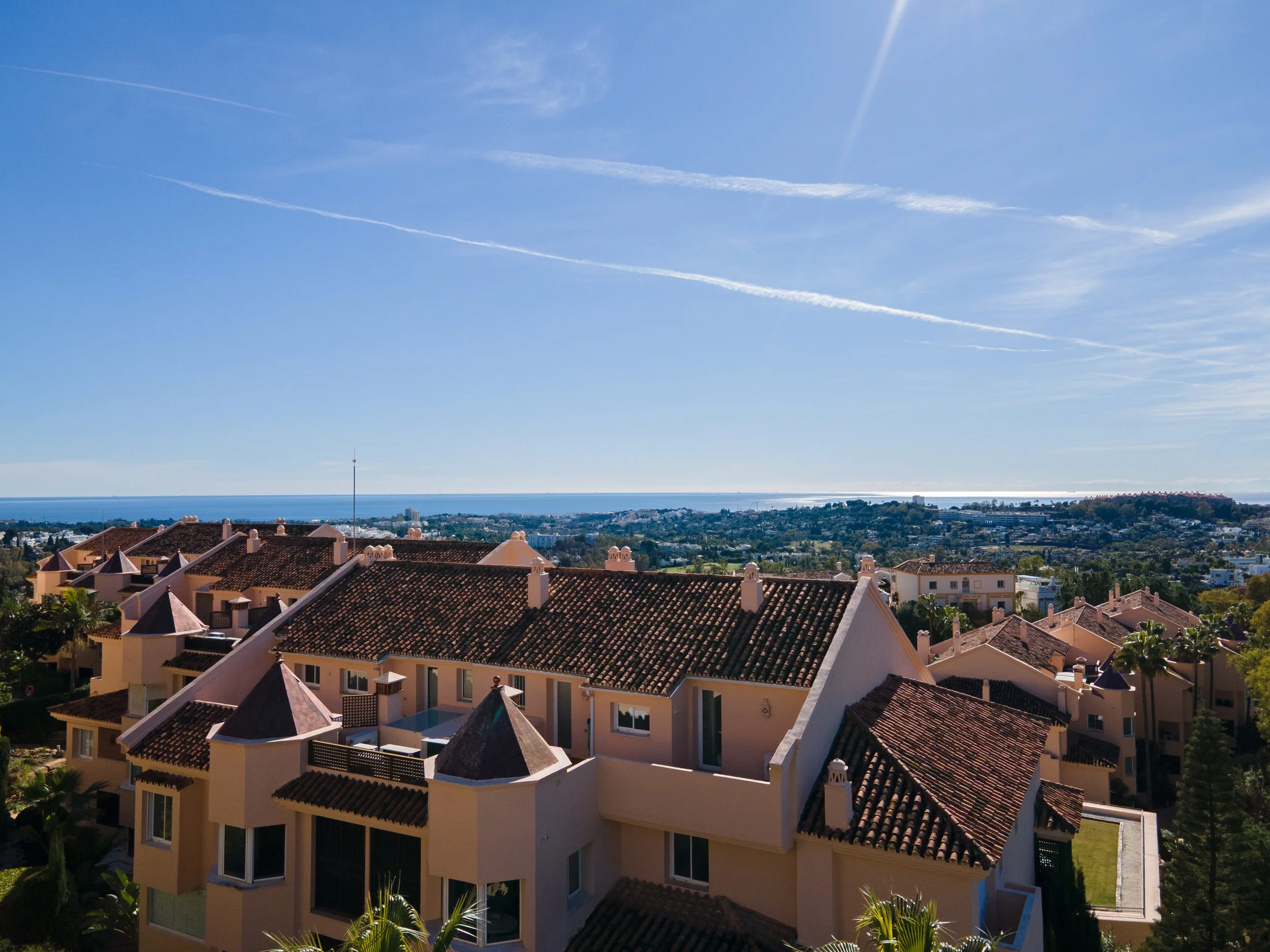 View of residential houses with red-tiled roofs and a distant ocean under a blue sky with a few contrail clouds.
