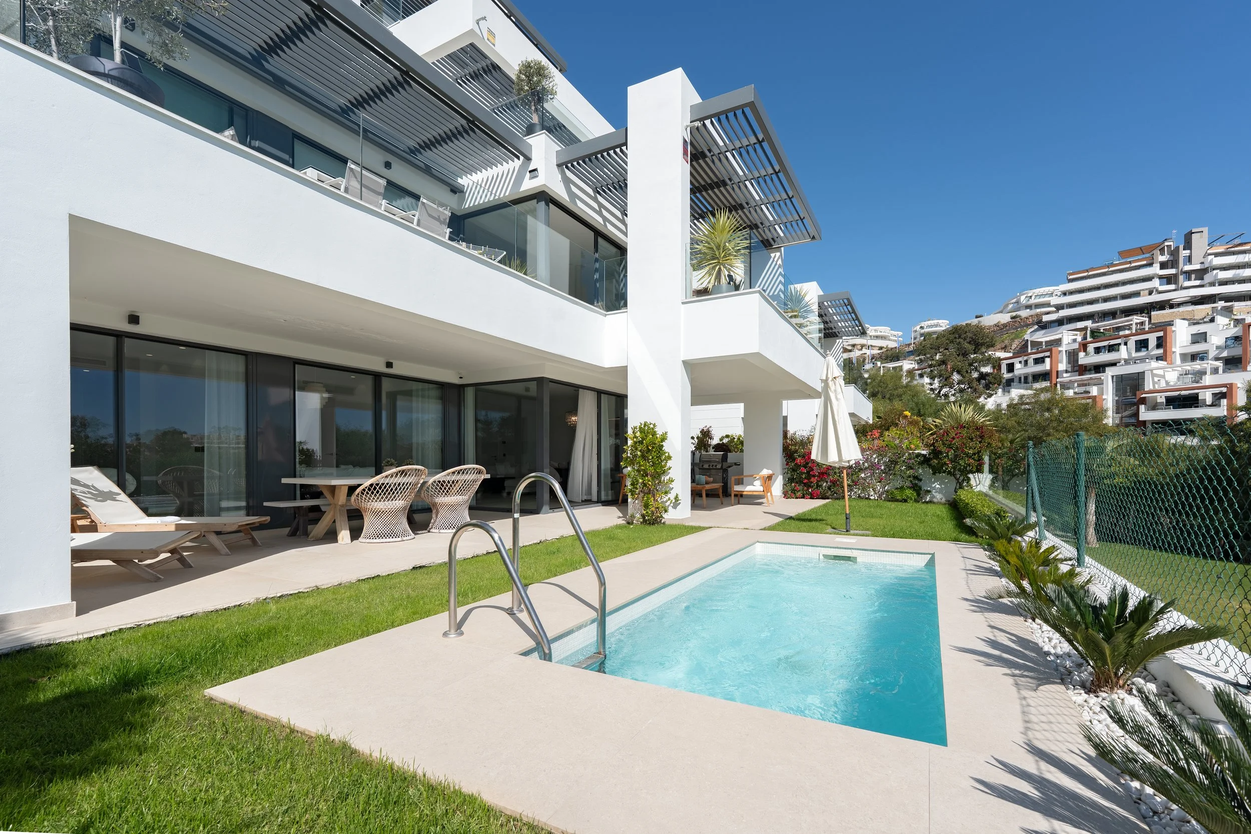 A modern white multi-story residential building with balconies, a small backyard with a swimming pool, outdoor furniture, and lush greenery, with hilly residential area in the background under a clear blue sky.