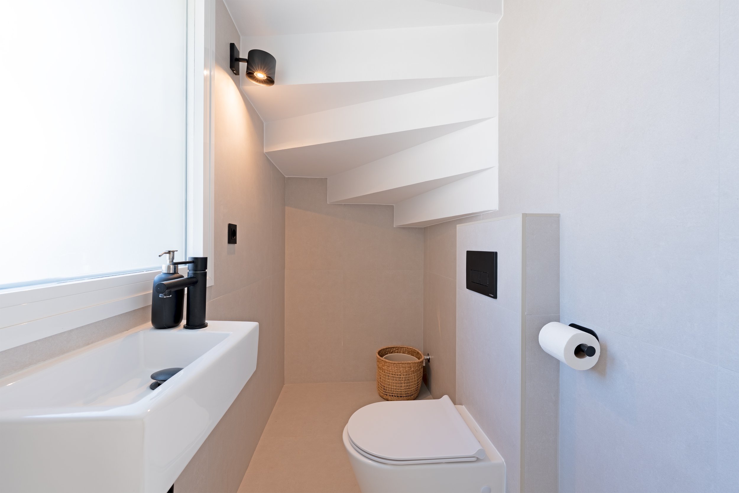 Small modern bathroom with a white sink, black faucet, wall-mounted toilet, toilet paper roll, small window, wall light, basket, and white staircase leading upwards.