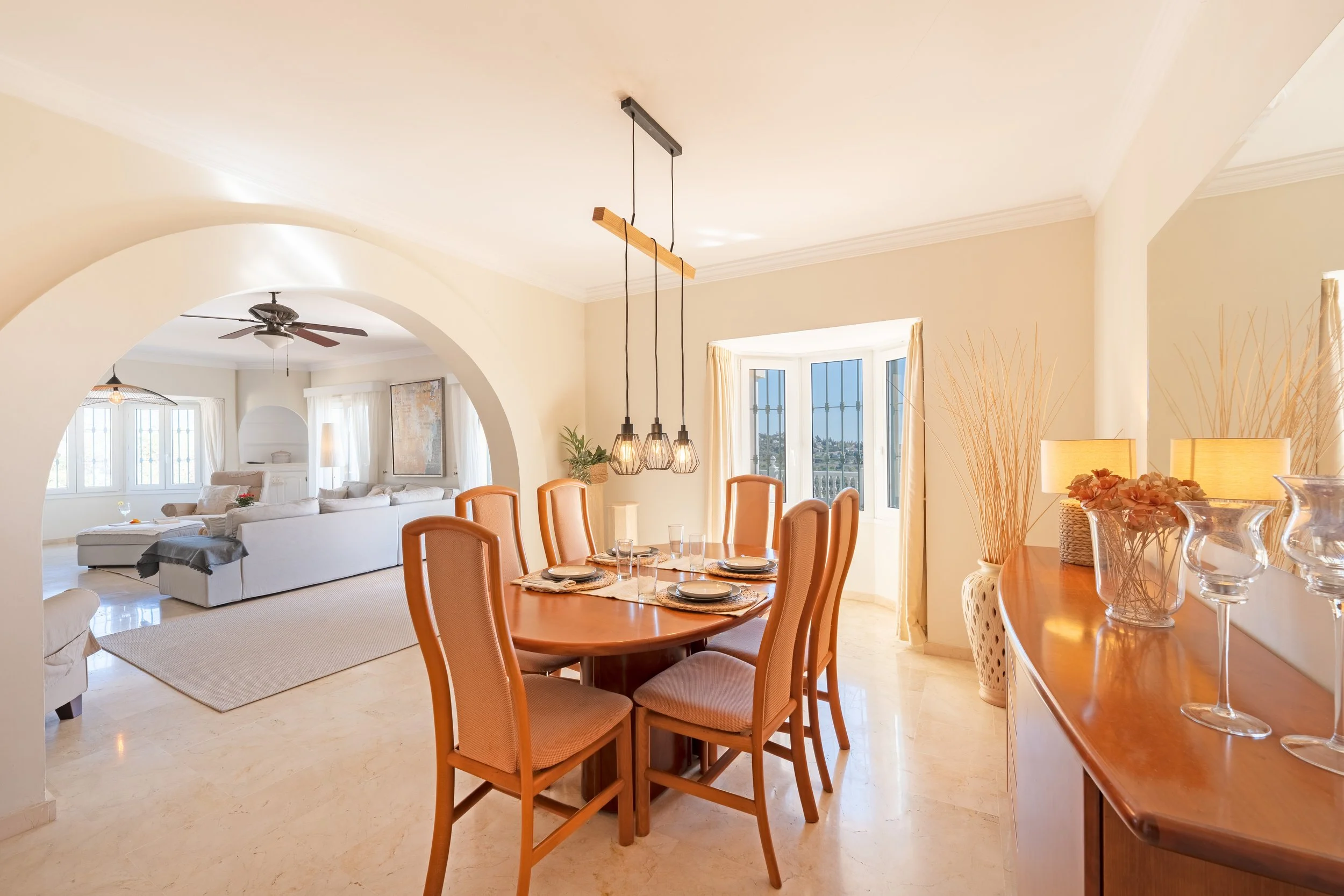 A dining room with a wooden table and six beige upholstered chairs, pendant lights hanging above, a built-in window with cream curtains, and a sideboard with vases and lamps, leading to a living room with sofas and a ceiling fan.