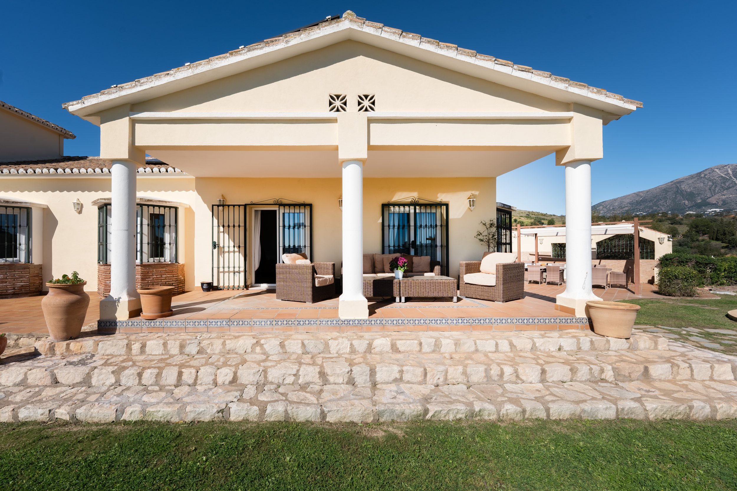 Front view of a house with a covered patio, outdoor seating, and potted plants, with a mountain and clear blue sky in the background.