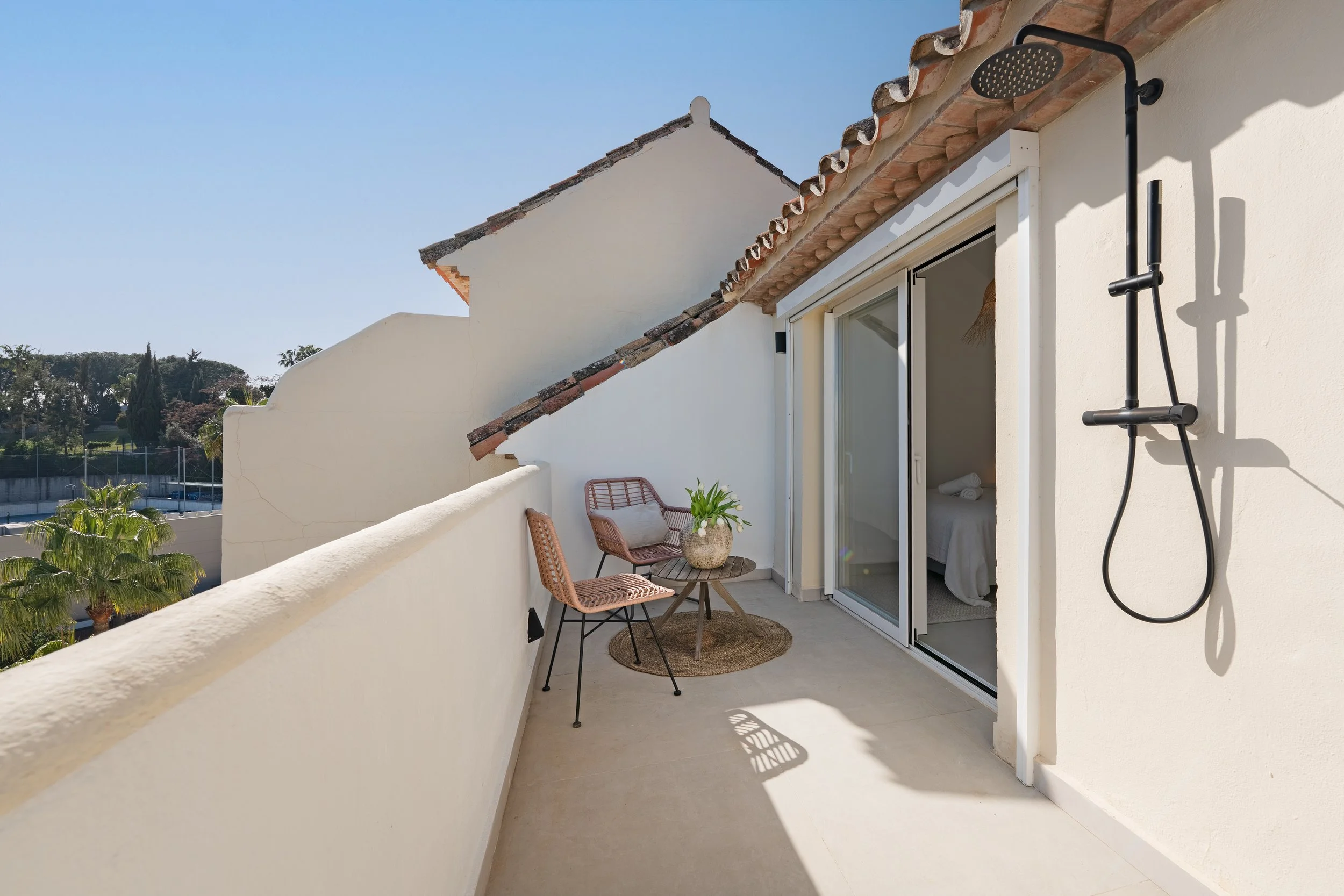 Balcony with two wicker chairs and a small round table with a potted plant, sliding glass doors leading into bedroom, outdoor shower on white wall, overlooking trees and tennis courts in the background.