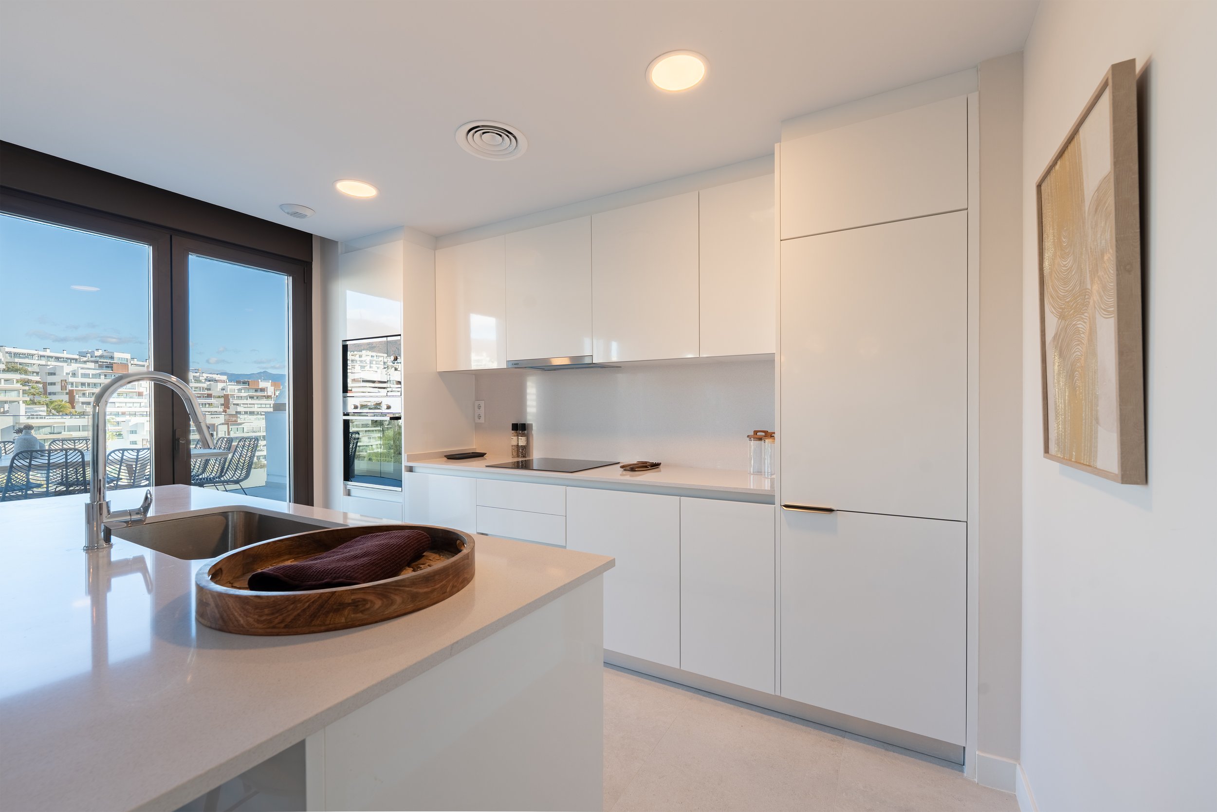 Modern, white kitchen with large glass sliding doors, a wooden tray with a dark cloth on the island, and minimal decor.